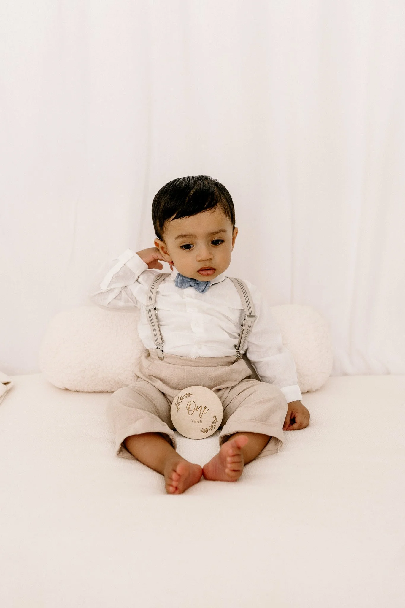 A young boy sitting on a white bed, wearing a white shirt, beige pants with suspenders, and a blue bow tie. There is a round sign in front of him that reads 'One Year.'