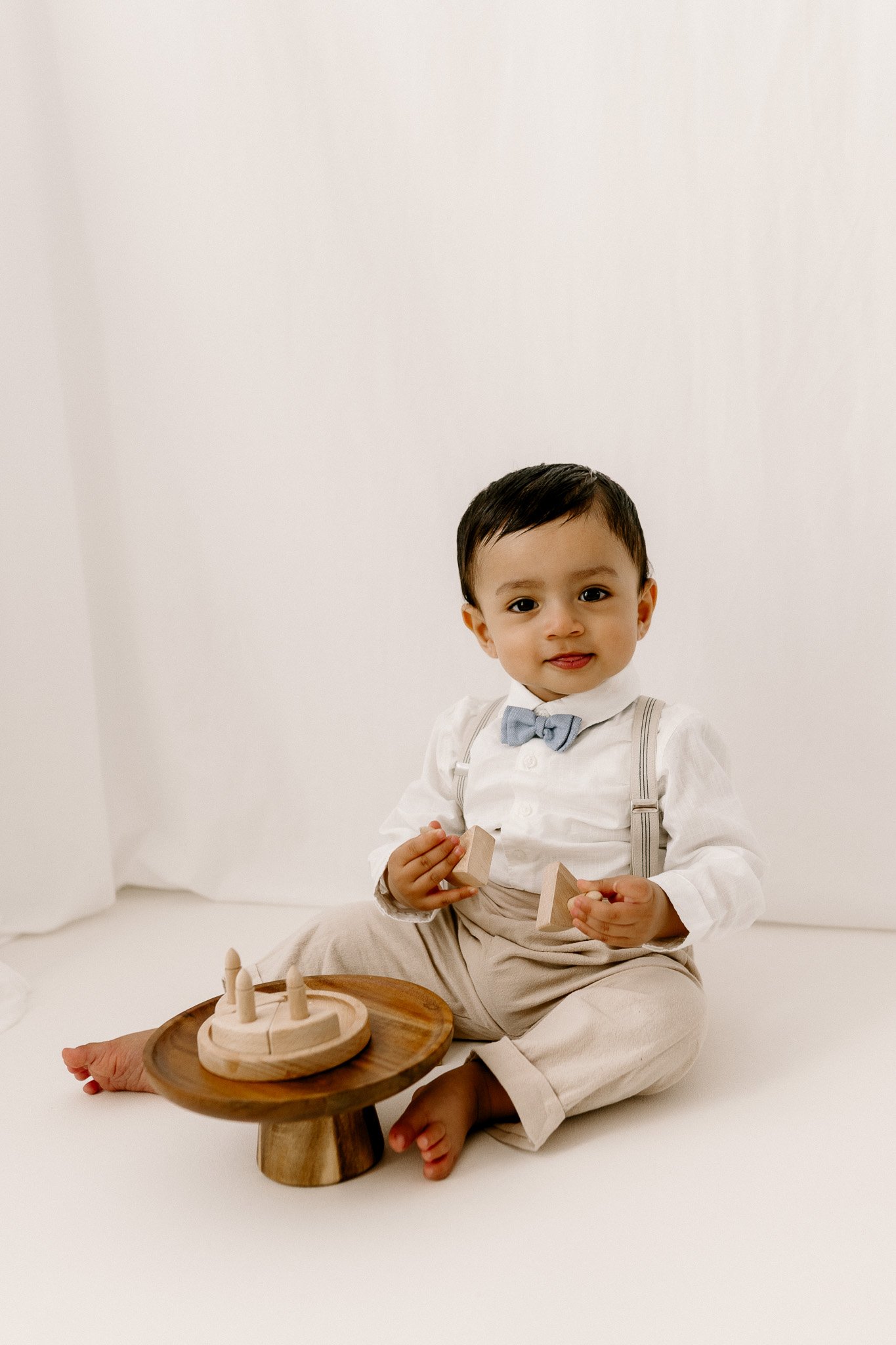 A young child dressed in a white shirt, beige trousers, a gray bow tie, and suspenders, sitting on the floor with a wooden toy and blocks, against a plain white background.