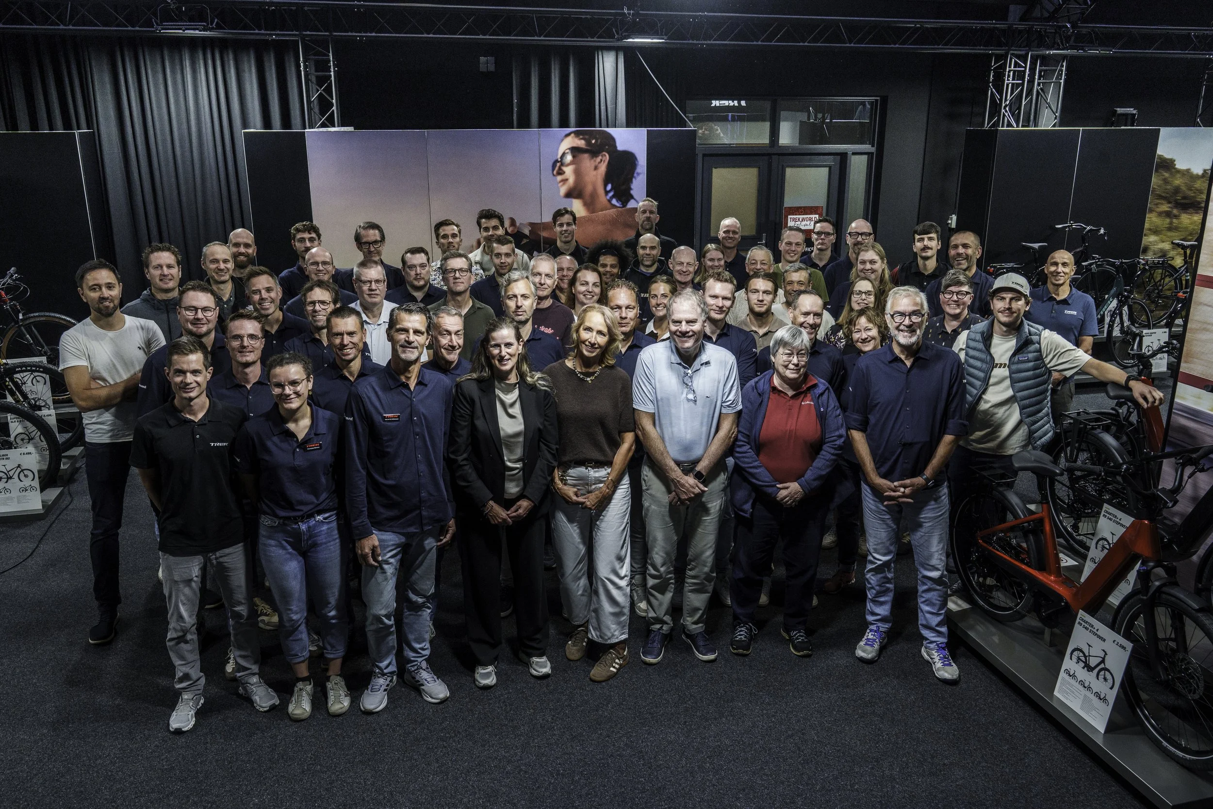 A large group of people is gathered inside a bicycle store, posing for a group photo. The store features bicycles on display and a large screen behind the group showing a woman in sunglasses.