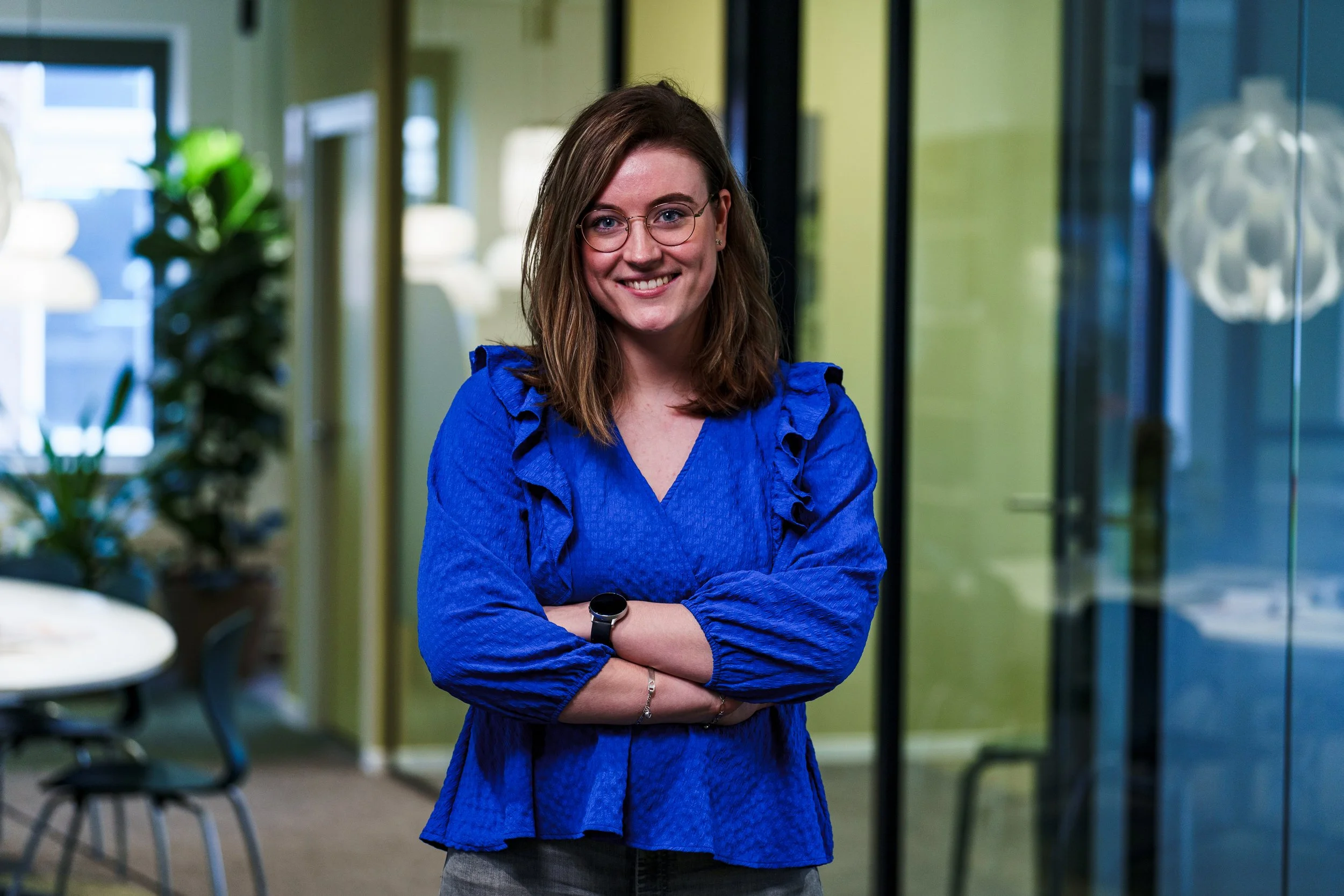 A woman with shoulder-length brown hair, glasses, and a blue blouse standing with crossed arms inside an office, smiling at the camera.