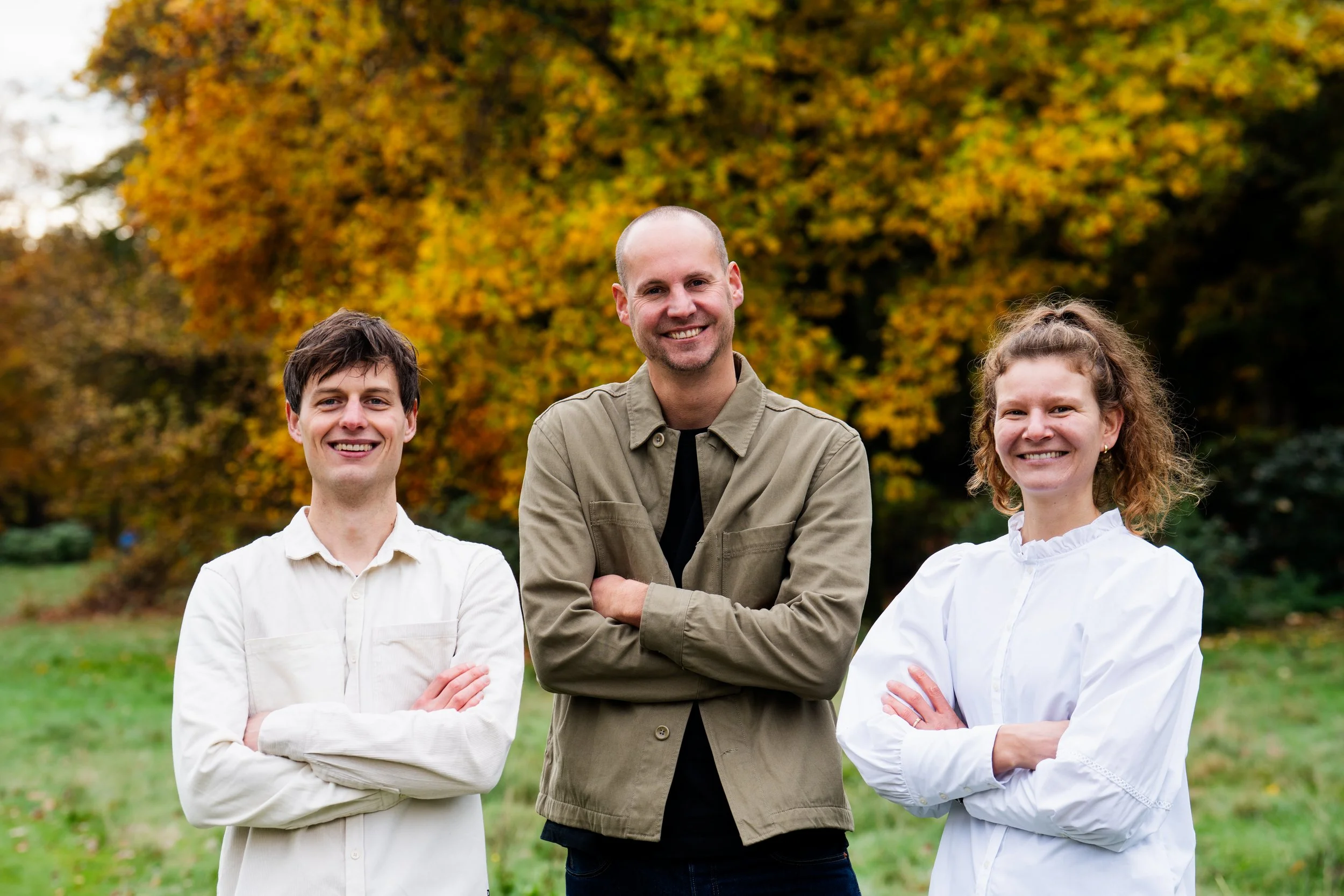 Three smiling people standing outdoors in a park with autumn-colored trees in the background.