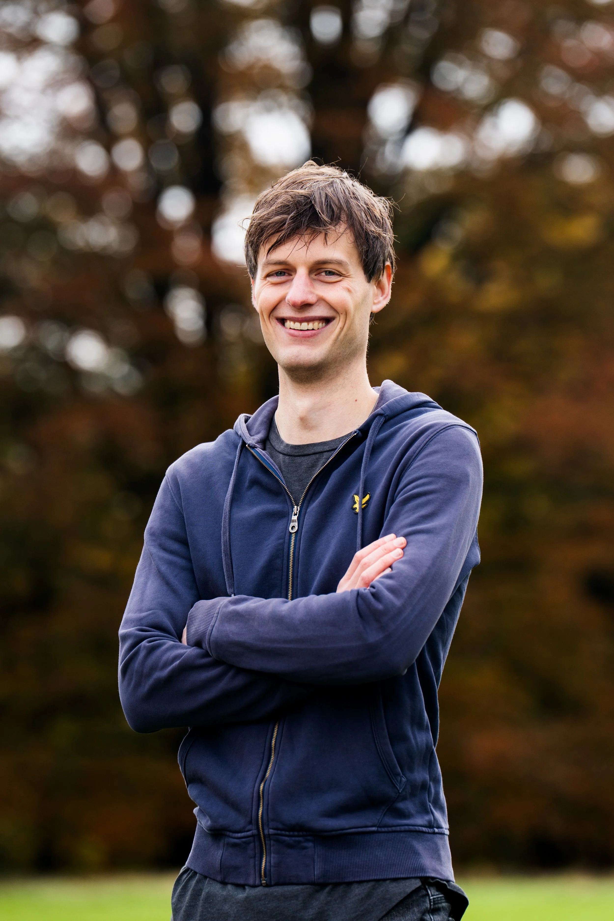 A young man with brown hair, smiling, wearing a navy zip-up hoodie and gray t-shirt, standing outdoors in front of trees with autumn leaves.