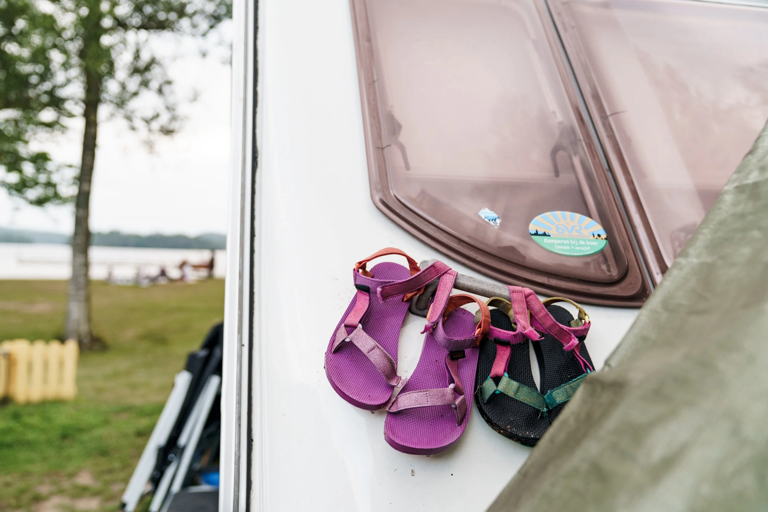 A pair of pink and black sandals hanging on the side of a white camper or trailer, with a window and outdoor park scene in the background.