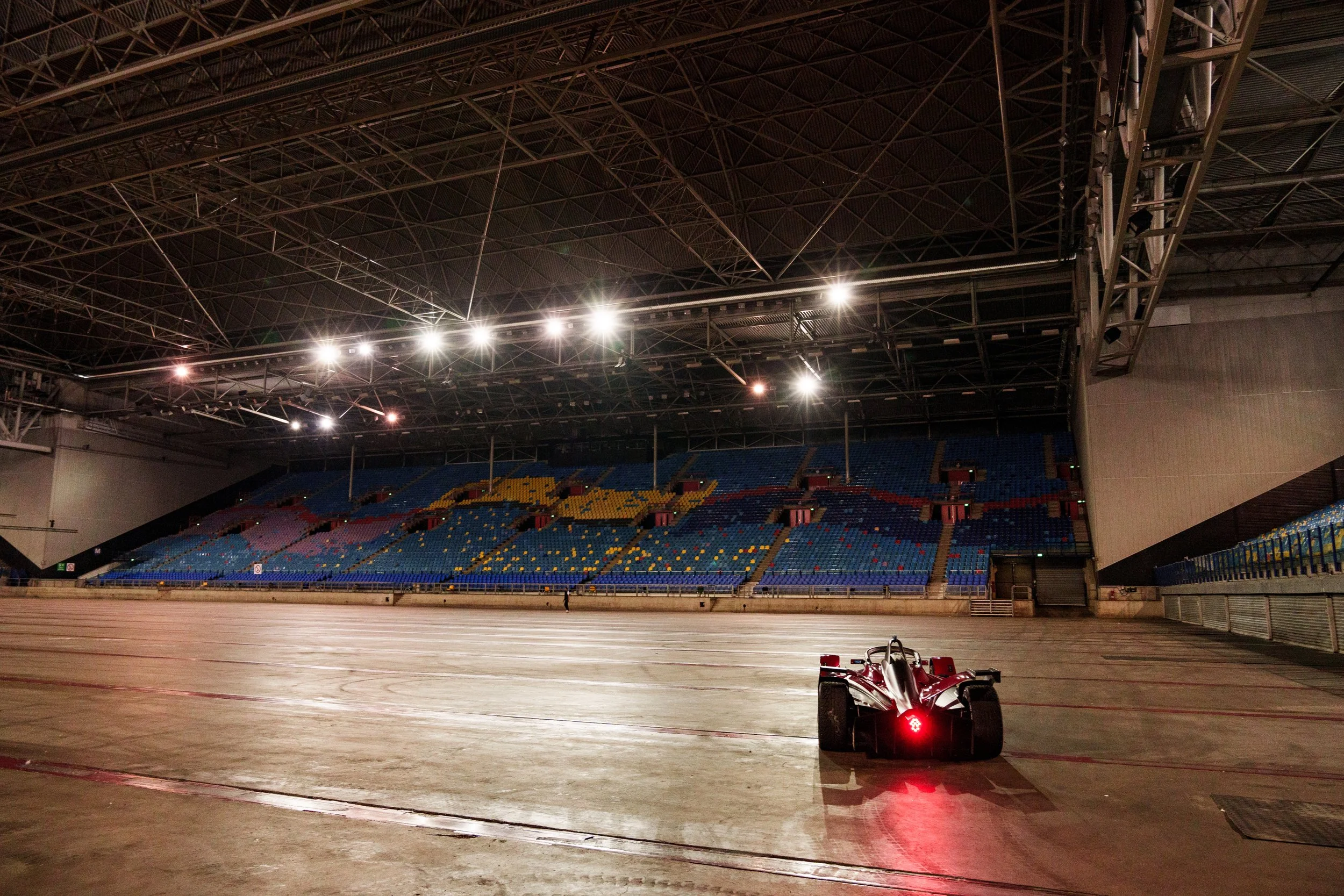 An empty indoor racing stadium with a single race car on the track, colorful seating in the stands, and bright lights illuminating the space.