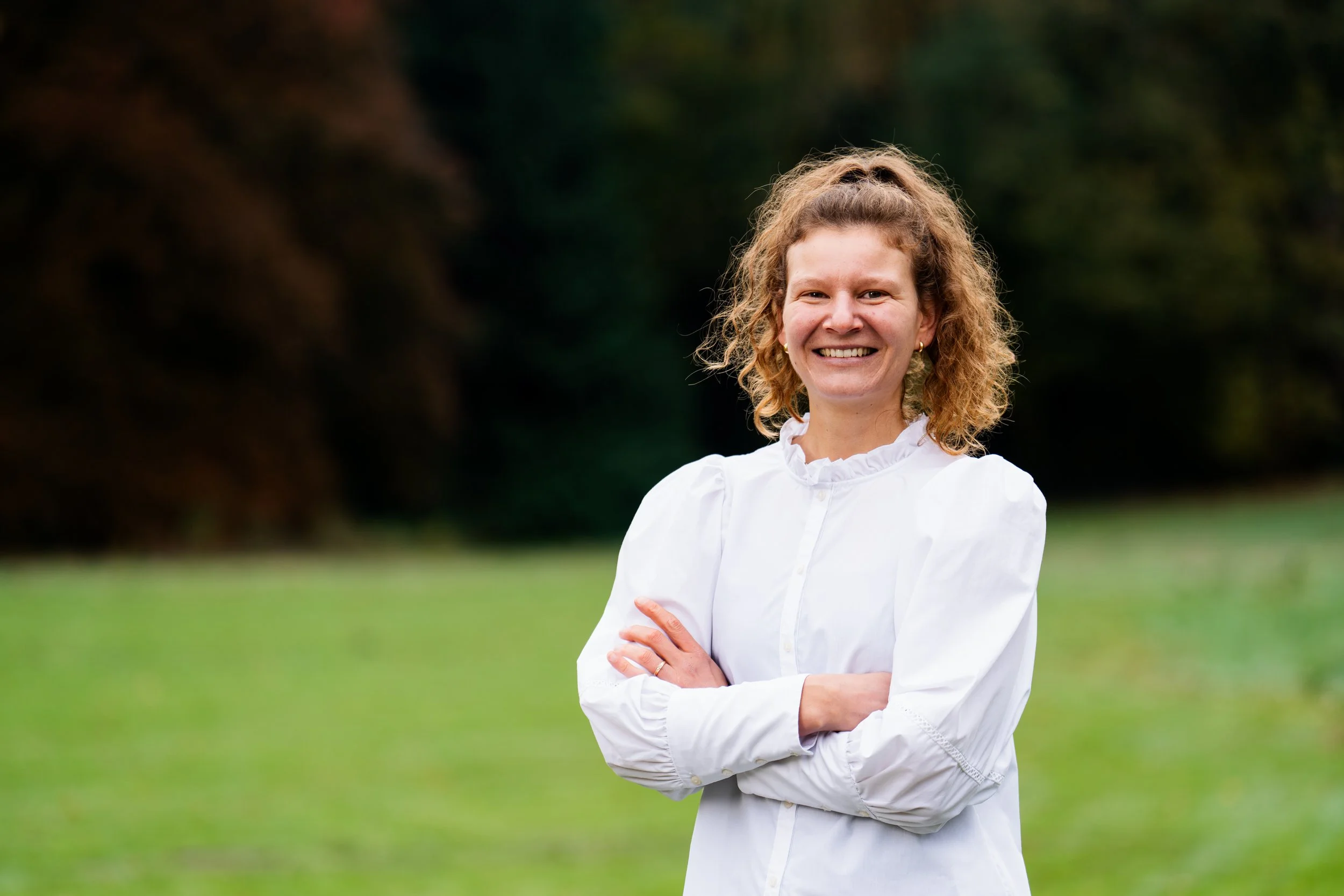 A woman with curly blonde hair smiling and crossing her arms outdoors in a green park.