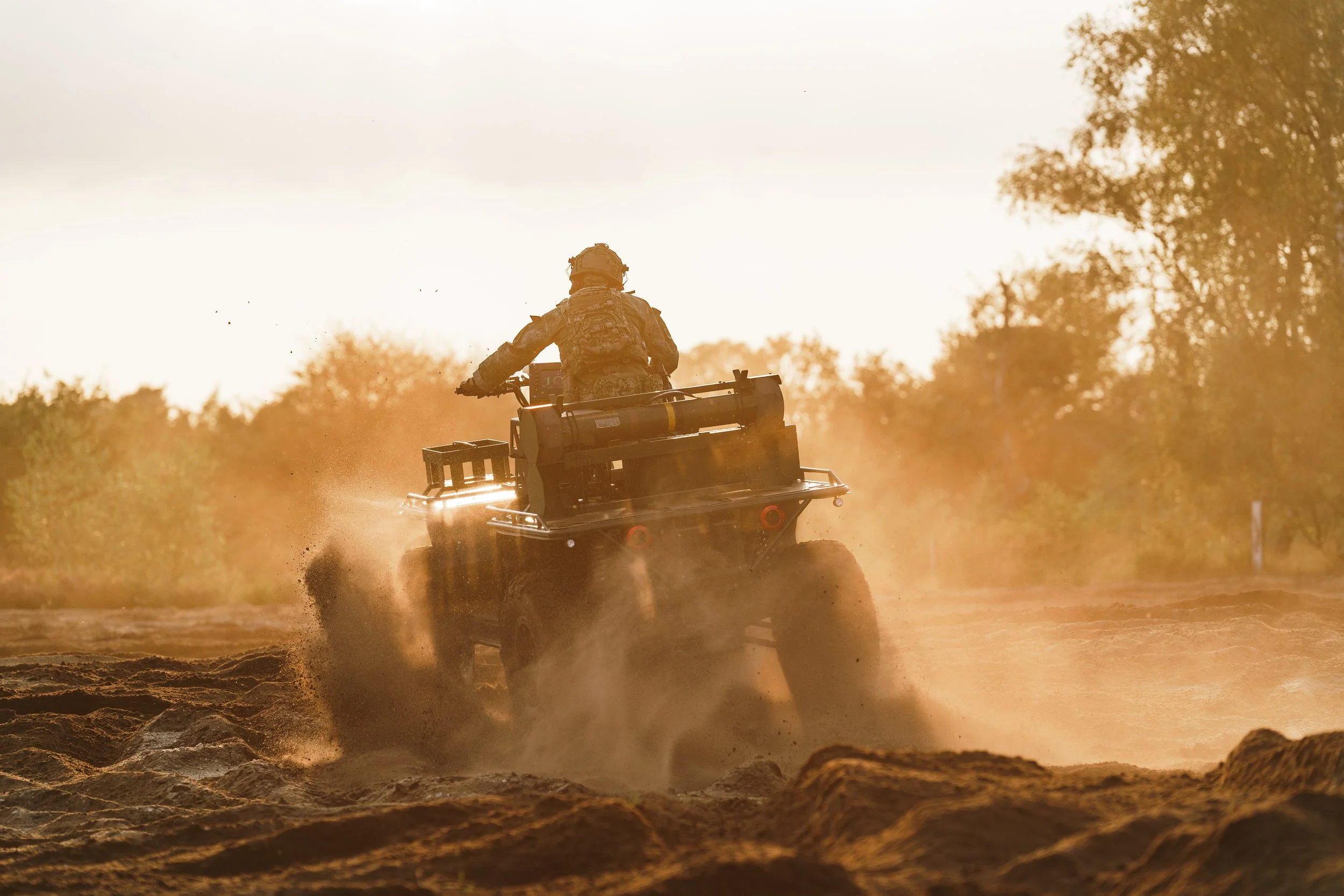 A soldier riding an all-terrain vehicle through a dirt field during sunset, kicking up dust and dirt.