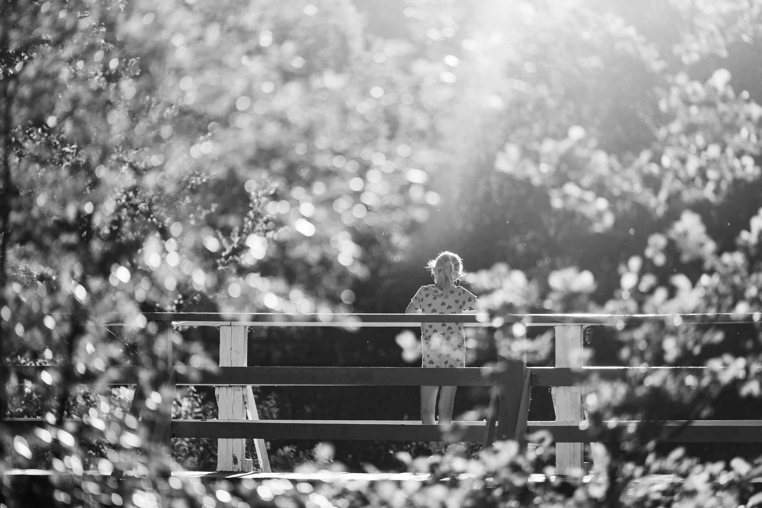 A young girl standing alone on a bridge with trees and foliage around her, illuminated by sunlight in black and white.