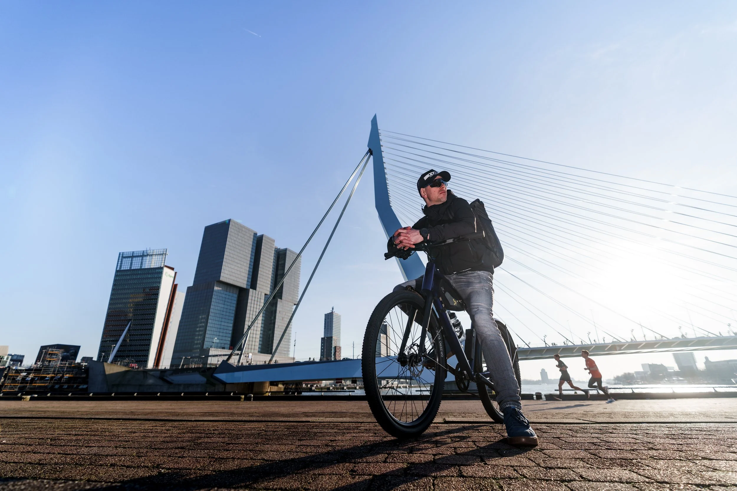 A man with sunglasses and a black backpack standing with a bicycle in front of a modern cityscape with tall buildings and a bridge during daytime.