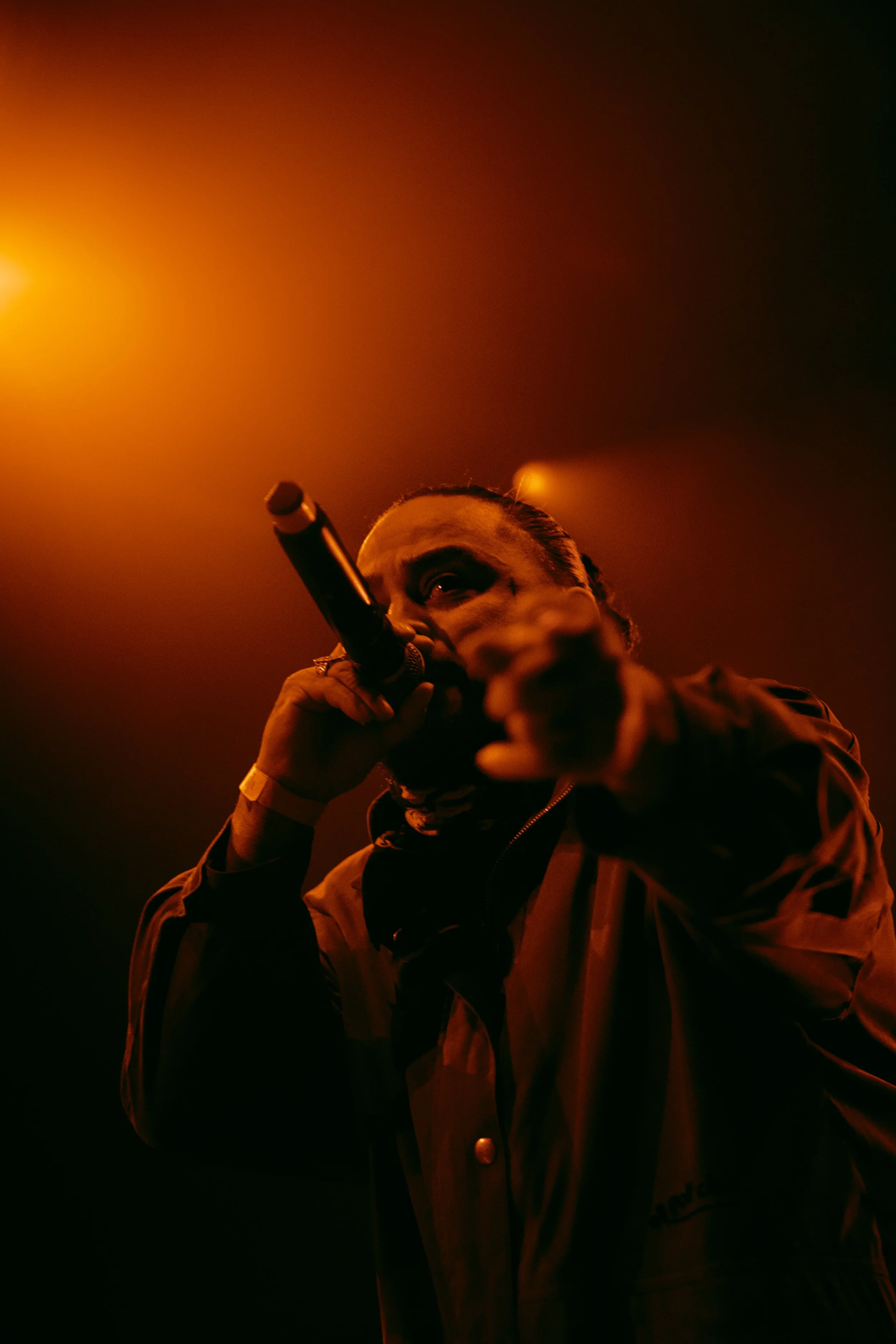 Performer with bracelet on wrist holding microphone, performing on stage with orange and black lighting.