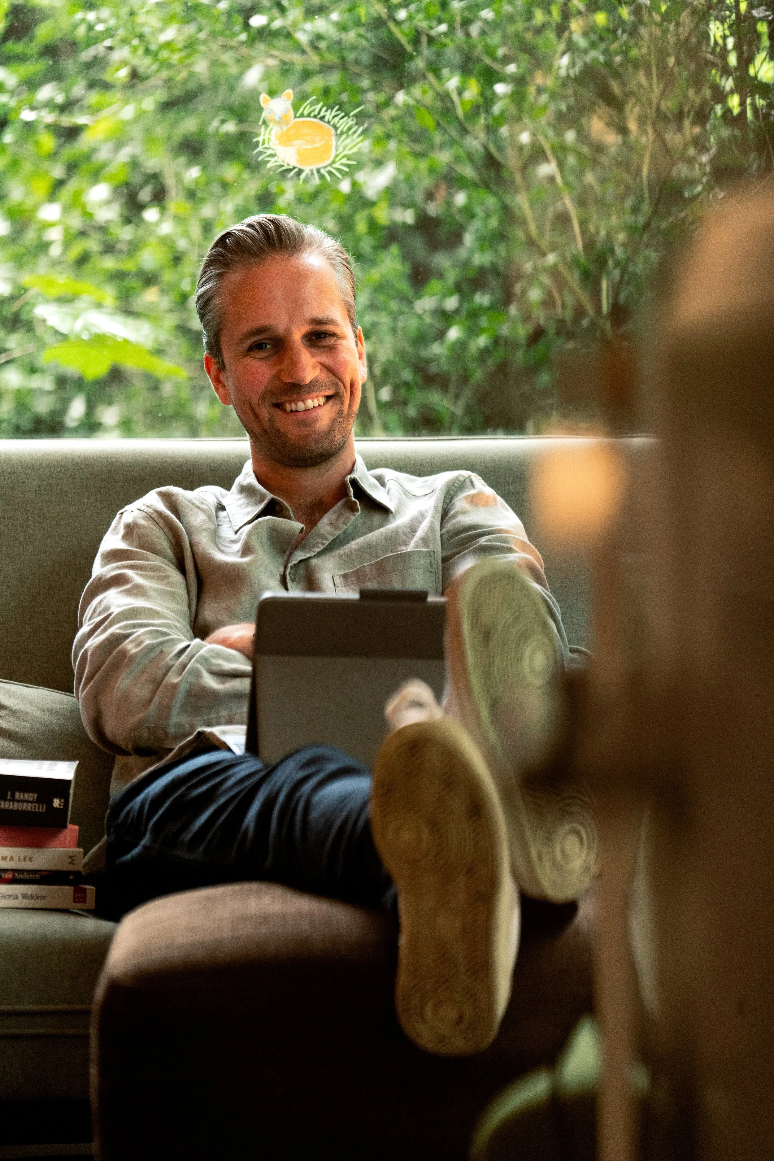A man with gray hair and a beard, smiling and sitting on a couch with his legs crossed, holding a tablet. There are books next to him and a large window with greenery in the background.