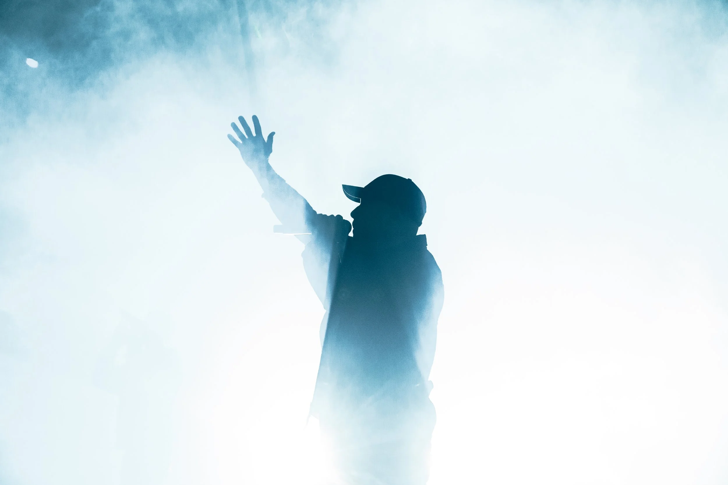 Silhouette of a person wearing a cap, raising their right hand amid fog or smoke, with a glow or light behind.