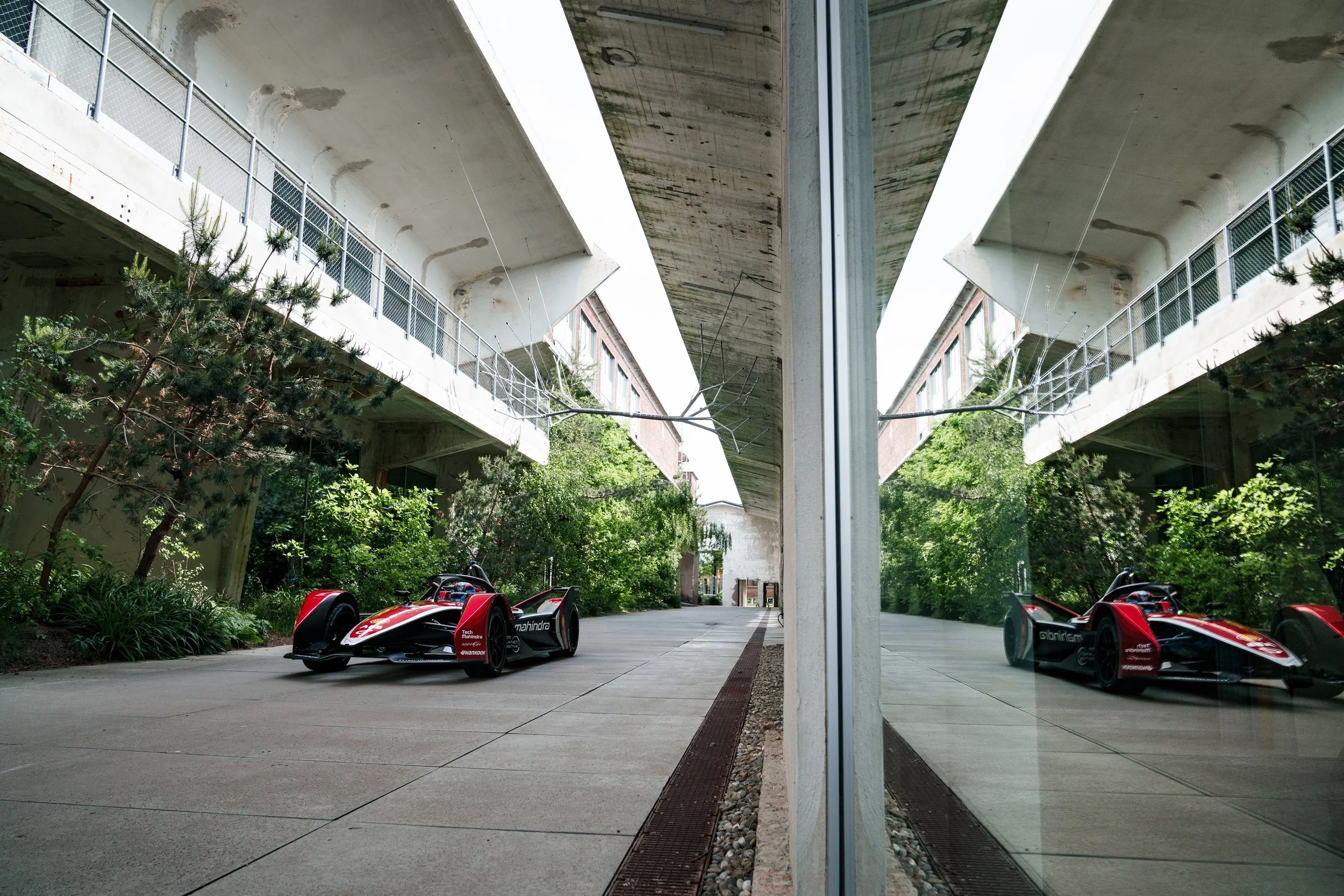 Reflected image of a red and black racing car parked on a concrete driveway, viewed through a glass window, with green trees and a concrete overpass in the background.