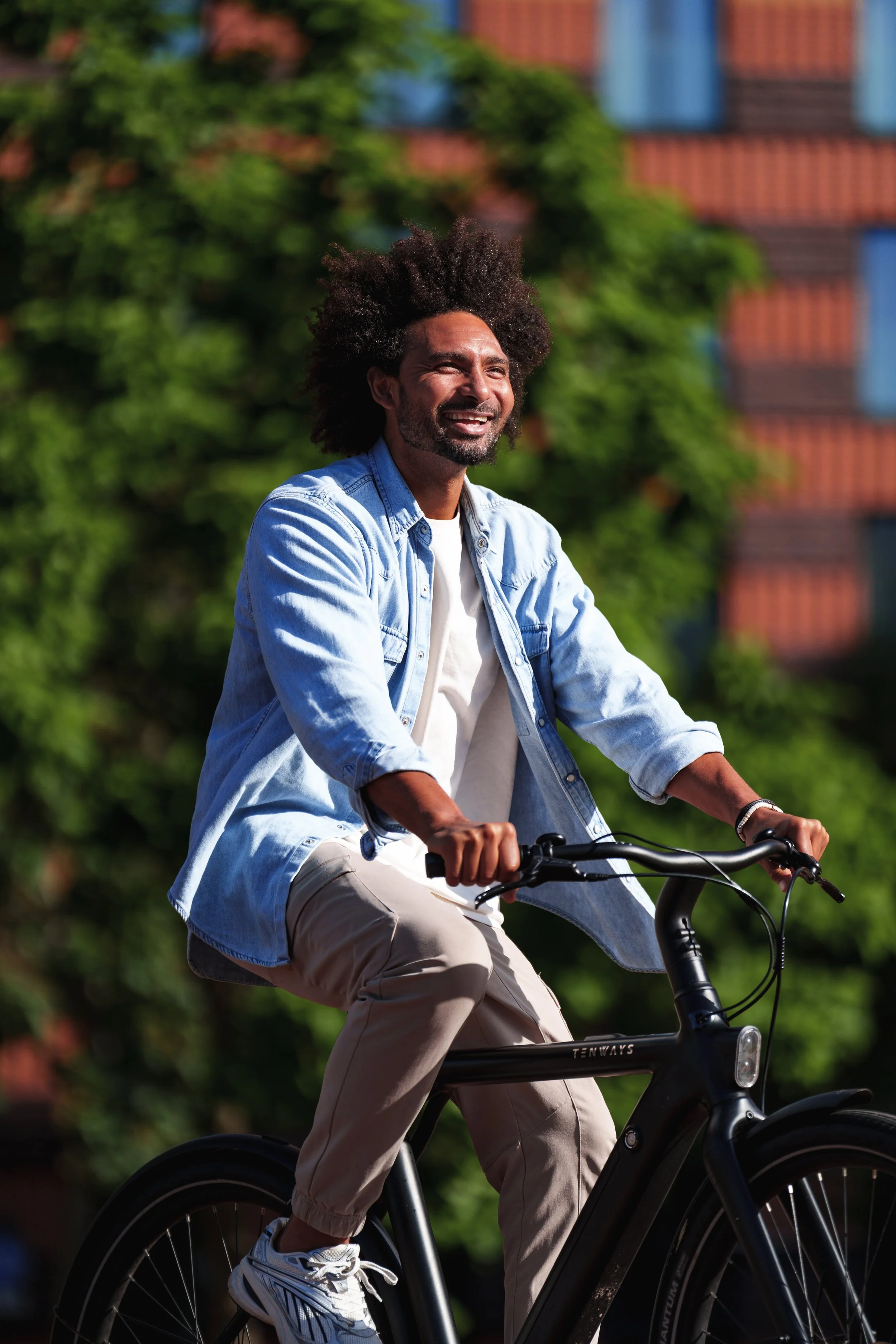 A man with curly hair riding a black bicycle outdoors, smiling with greenery and a building in the background.