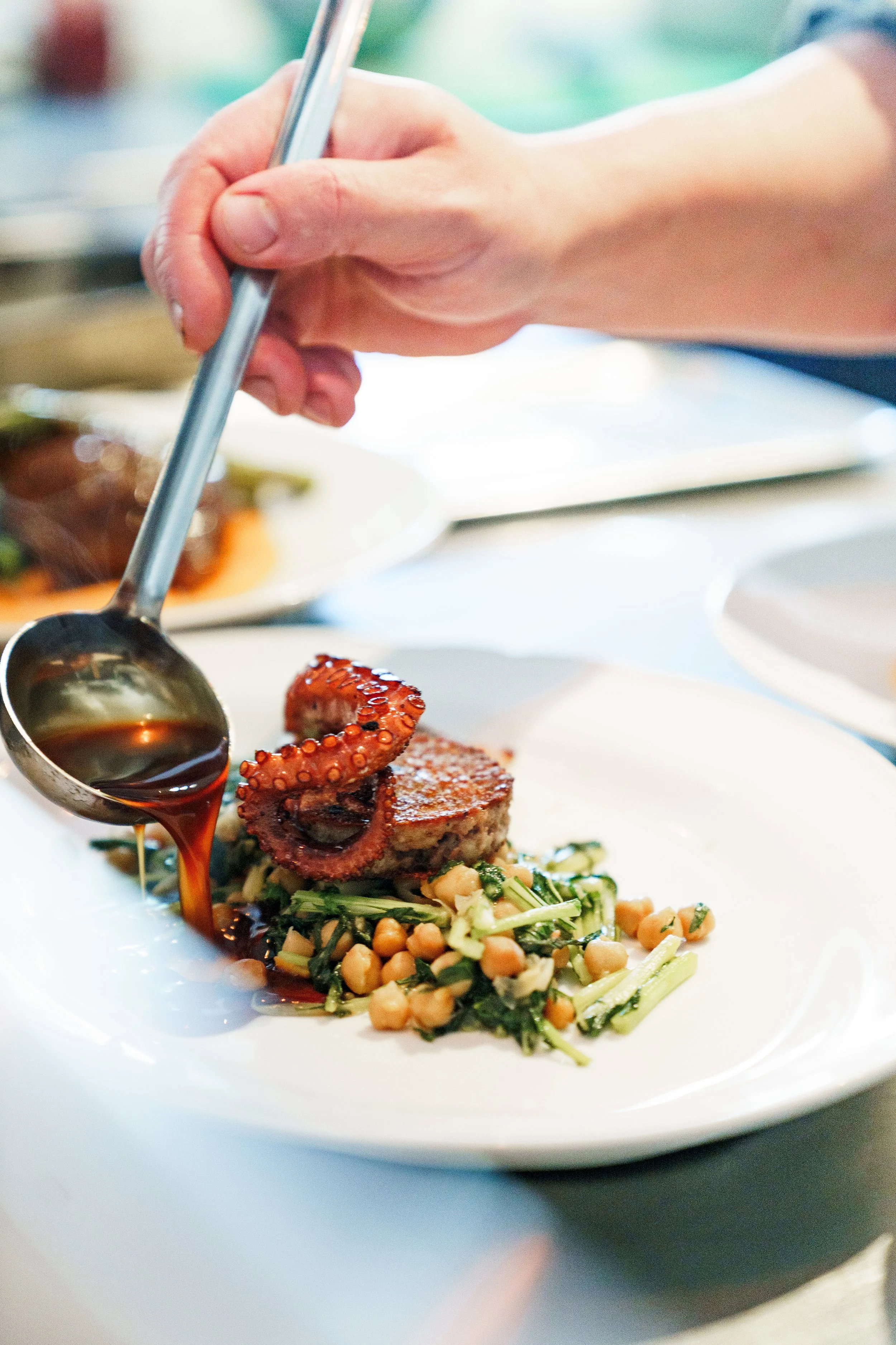 Person pouring sauce over a plated dish featuring grilled octopus, chickpeas, and greens.