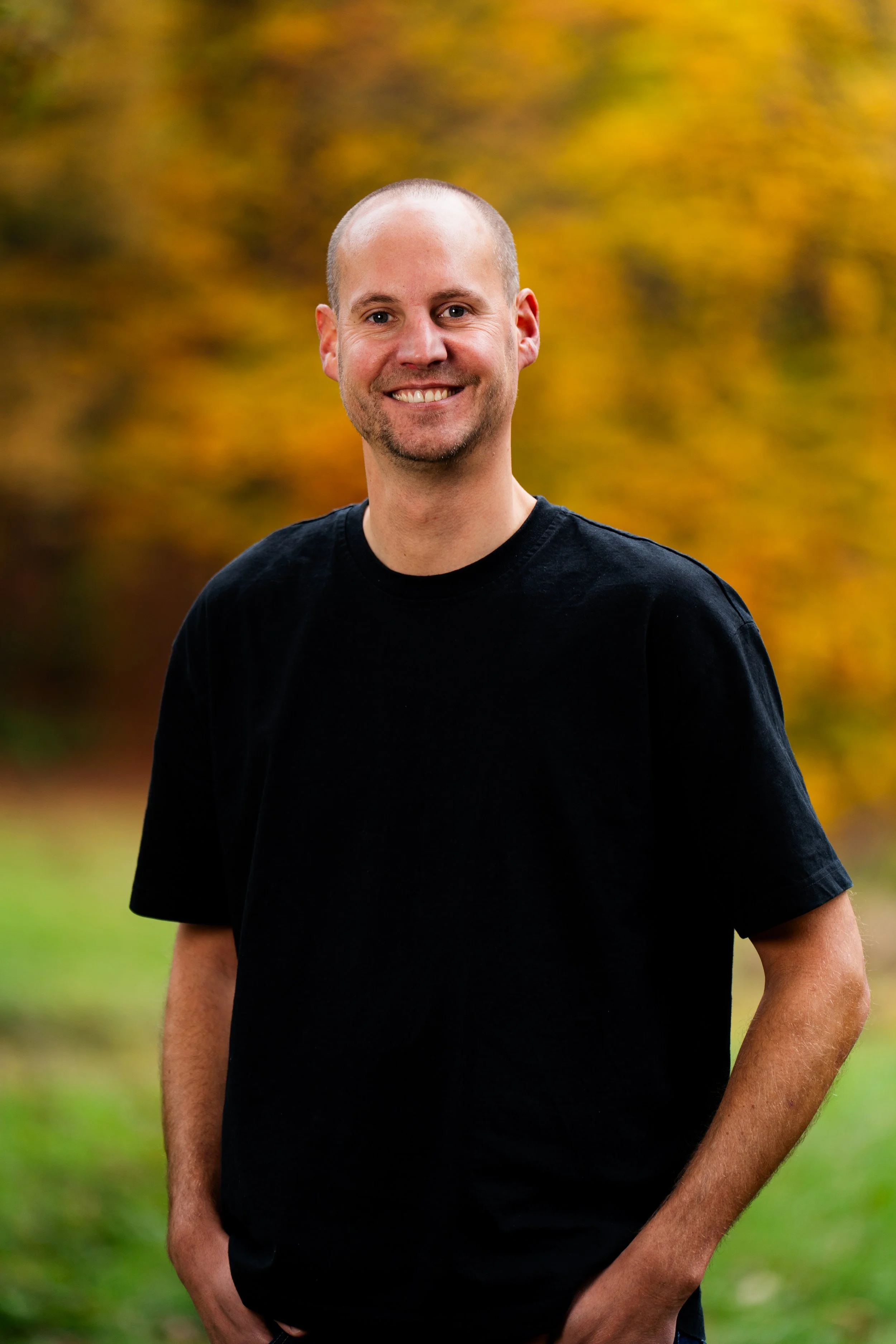 A smiling man with a shaved head and stubble, wearing a black t-shirt, standing outdoors in front of fall-colored trees.