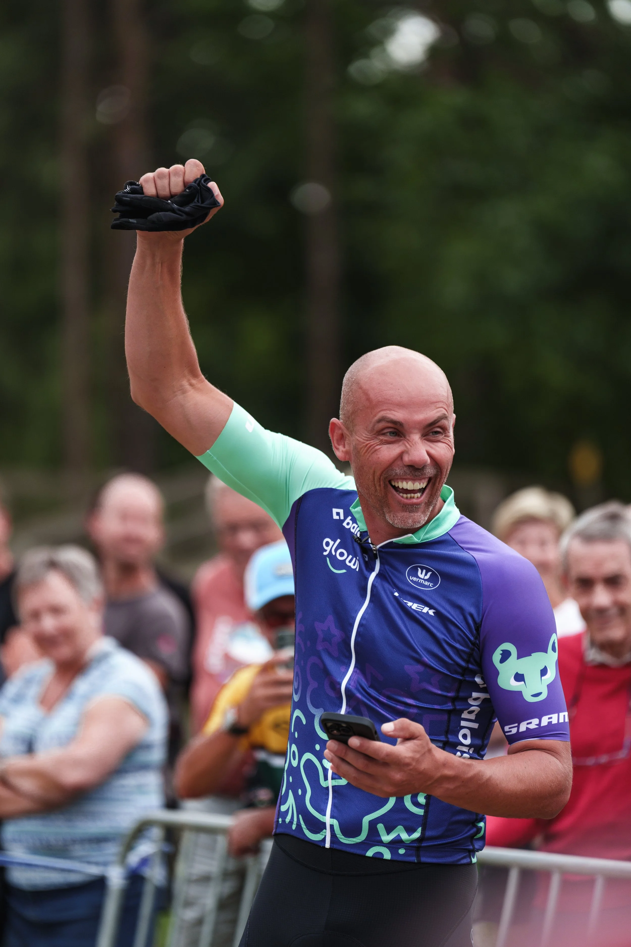 A smiling man in a colorful cycling jersey celebrates with a raised fist while holding a phone, surrounded by cheering spectators in an outdoor setting.