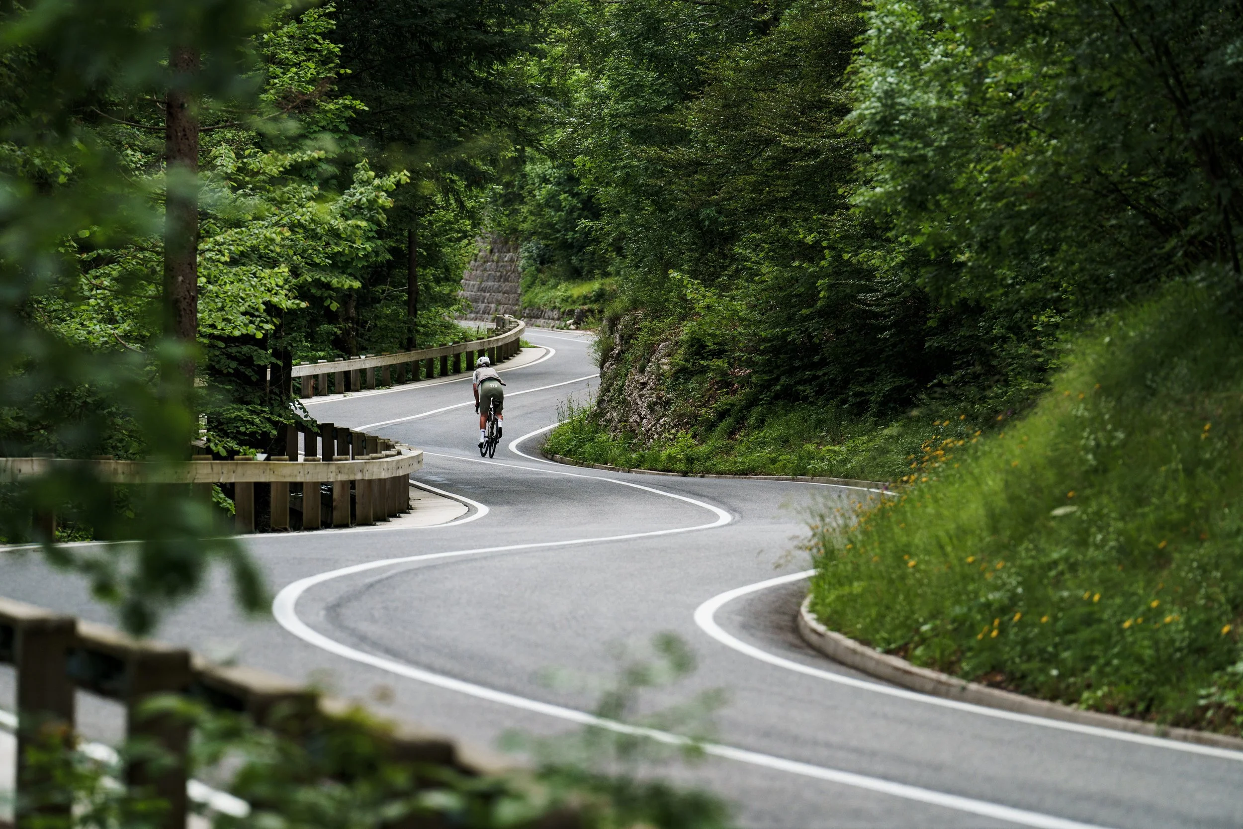 A winding mountain road through a forest with a cyclist riding uphill.