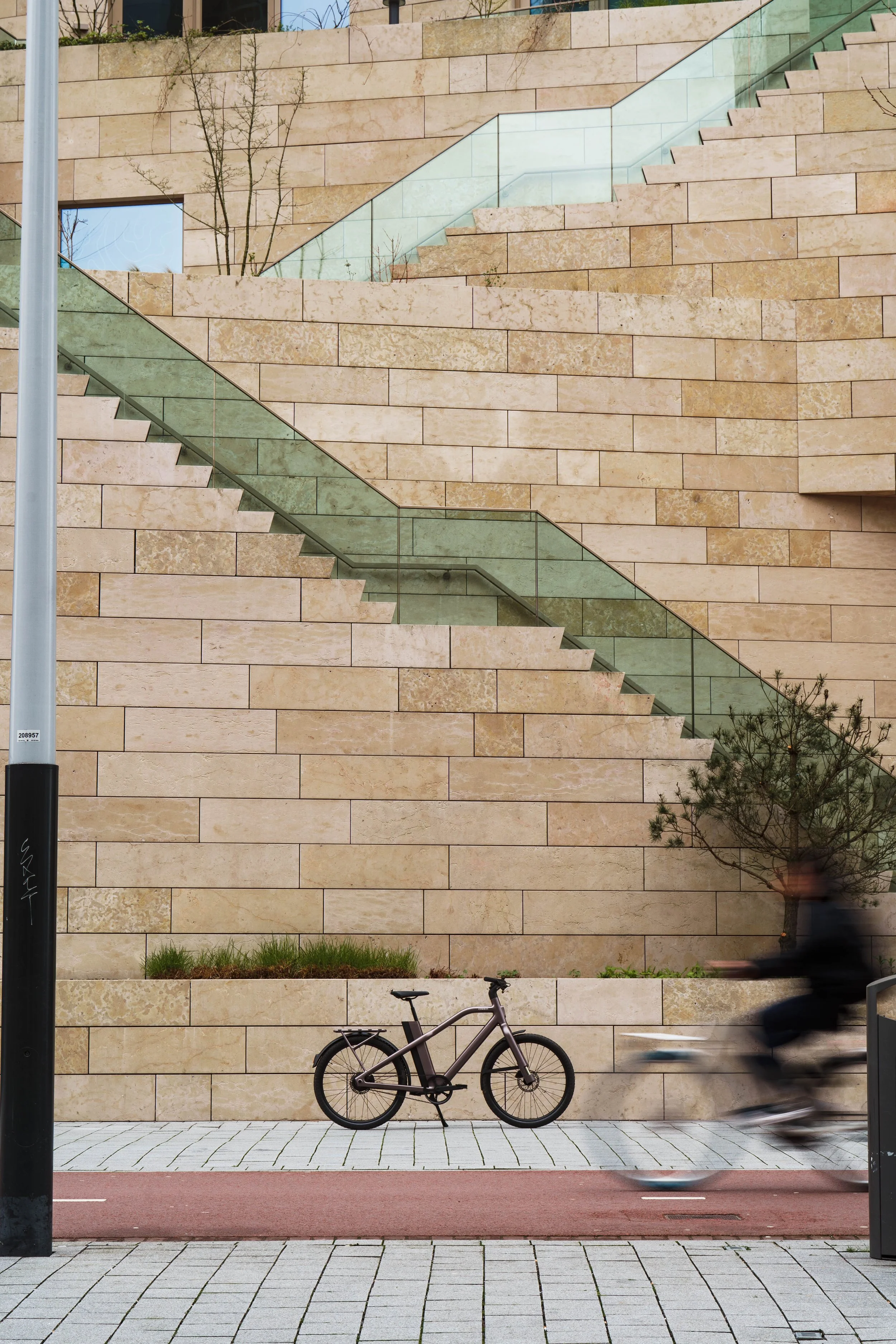 A modern urban scene with a beige stone wall and a glass stairway. In the foreground, a bicycle is parked on a paved sidewalk, with a blurred cyclist riding past. Small plants and a tree are visible against the wall.