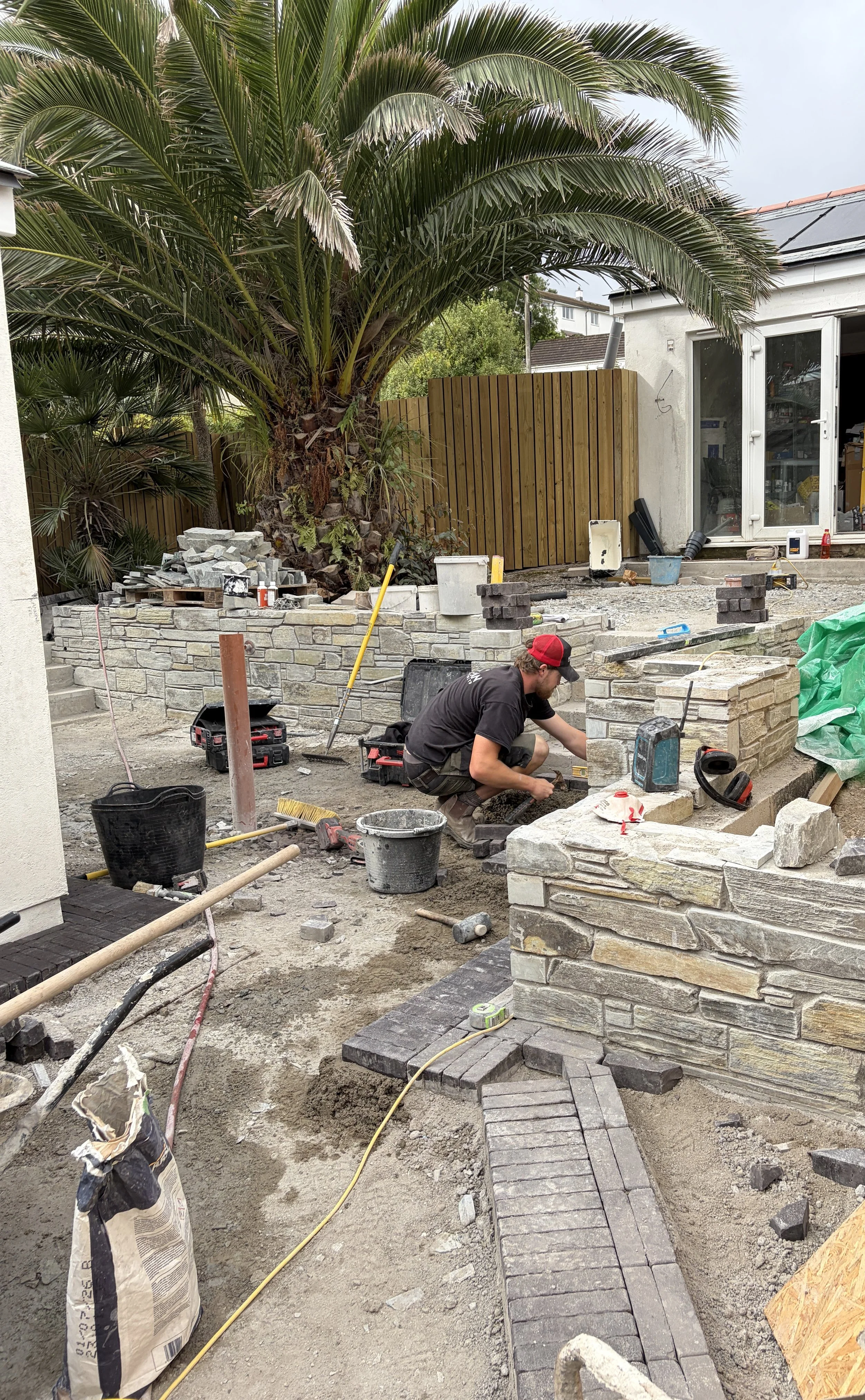A person working on a landscaping project outdoors, laying bricks to build a wall, with various tools and materials scattered around, including buckets, bags of mortar, and a pile of stones, adjacent to a house with a sliding glass door and a wooden fence.