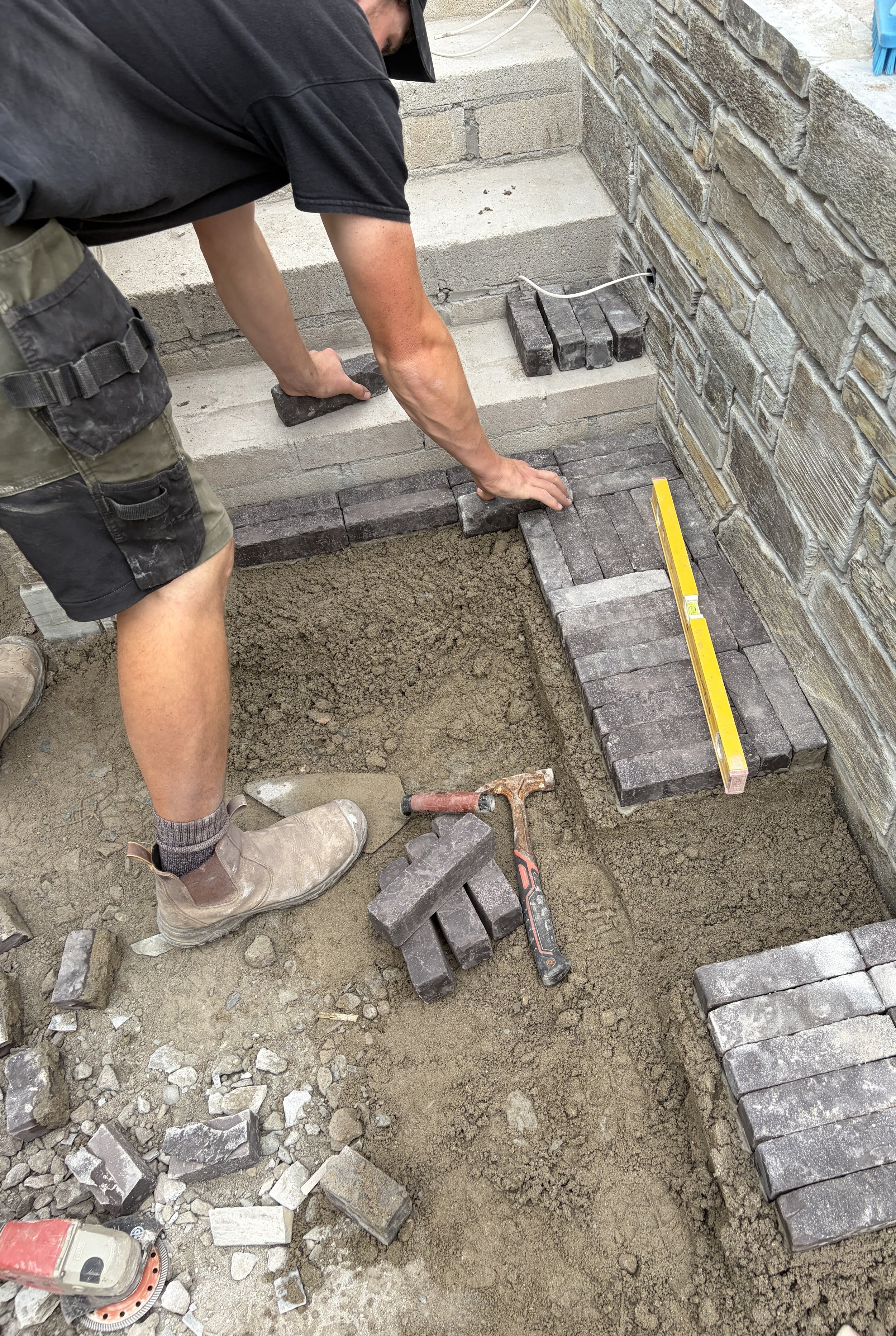 A construction worker laying bricks on a surface, using a level tool to ensure even placement. The worker is wearing work boots, shorts, a t-shirt, and a tool belt, and is working next to a brick wall.