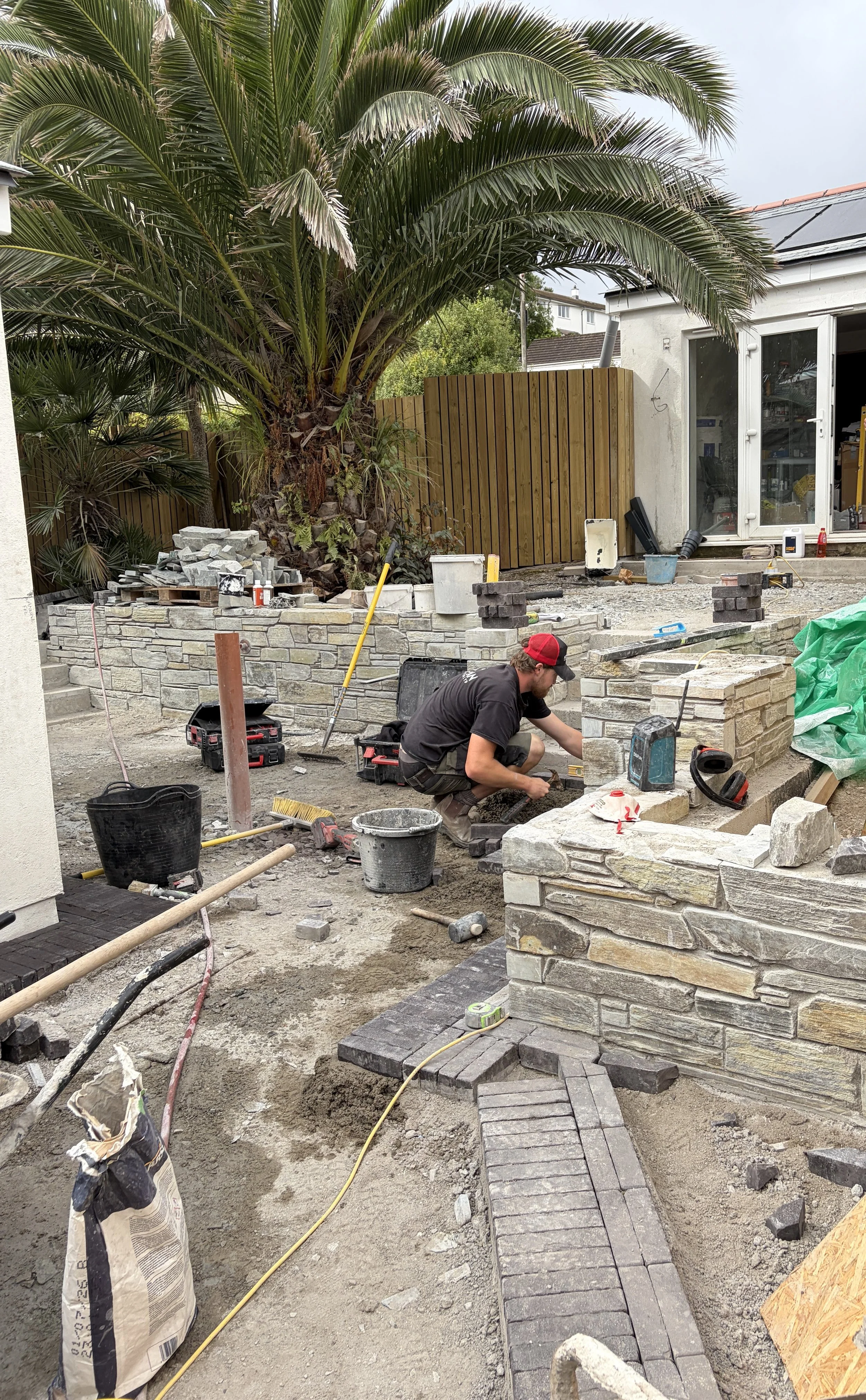 A person working on laying stone bricks for a garden or yard wall, with construction tools and materials around, in a backyard with trees and a house in the background.
