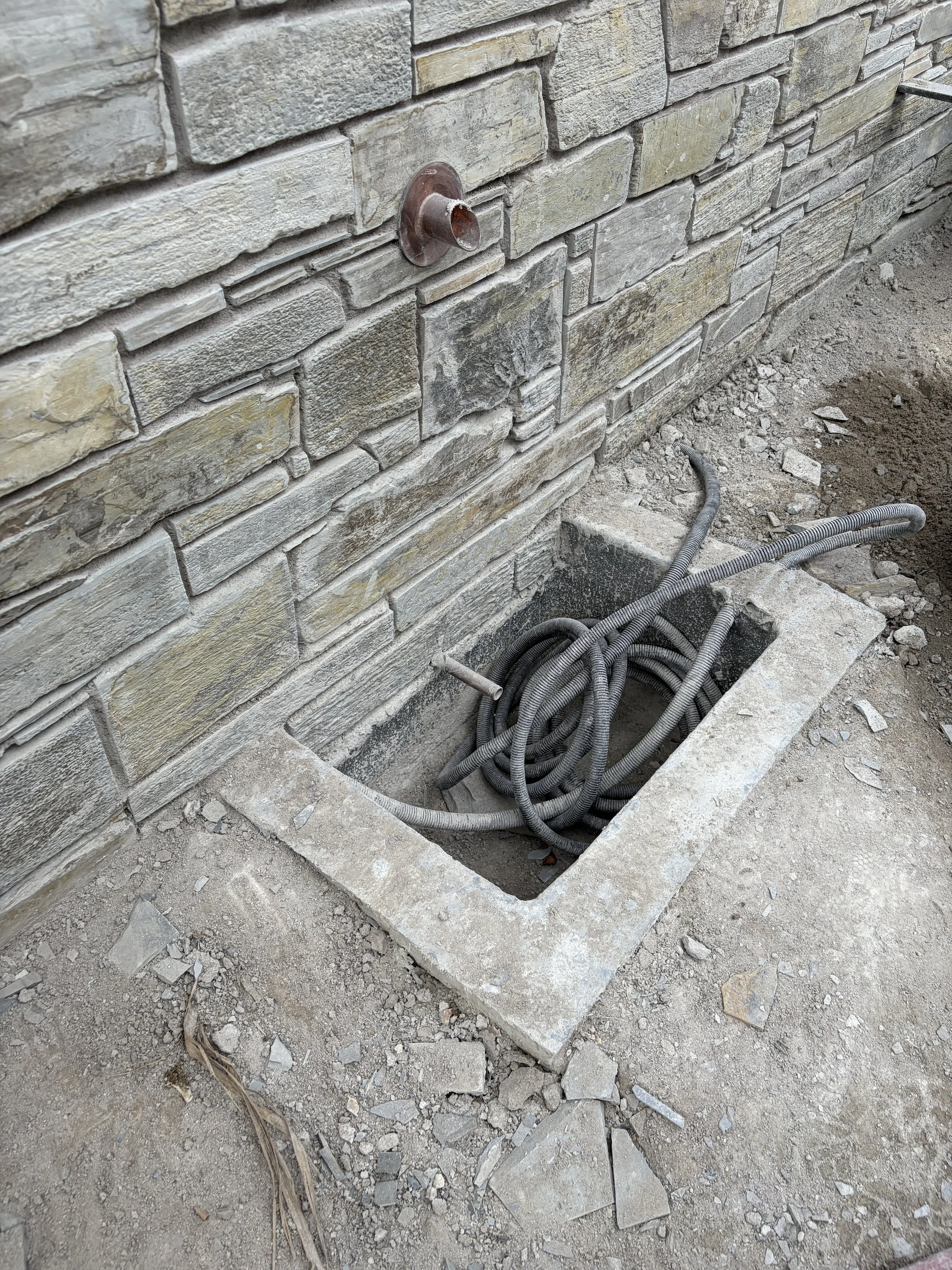 Close-up of a construction site with a brick wall and an open square hole in the concrete floor containing coiled electrical wires.