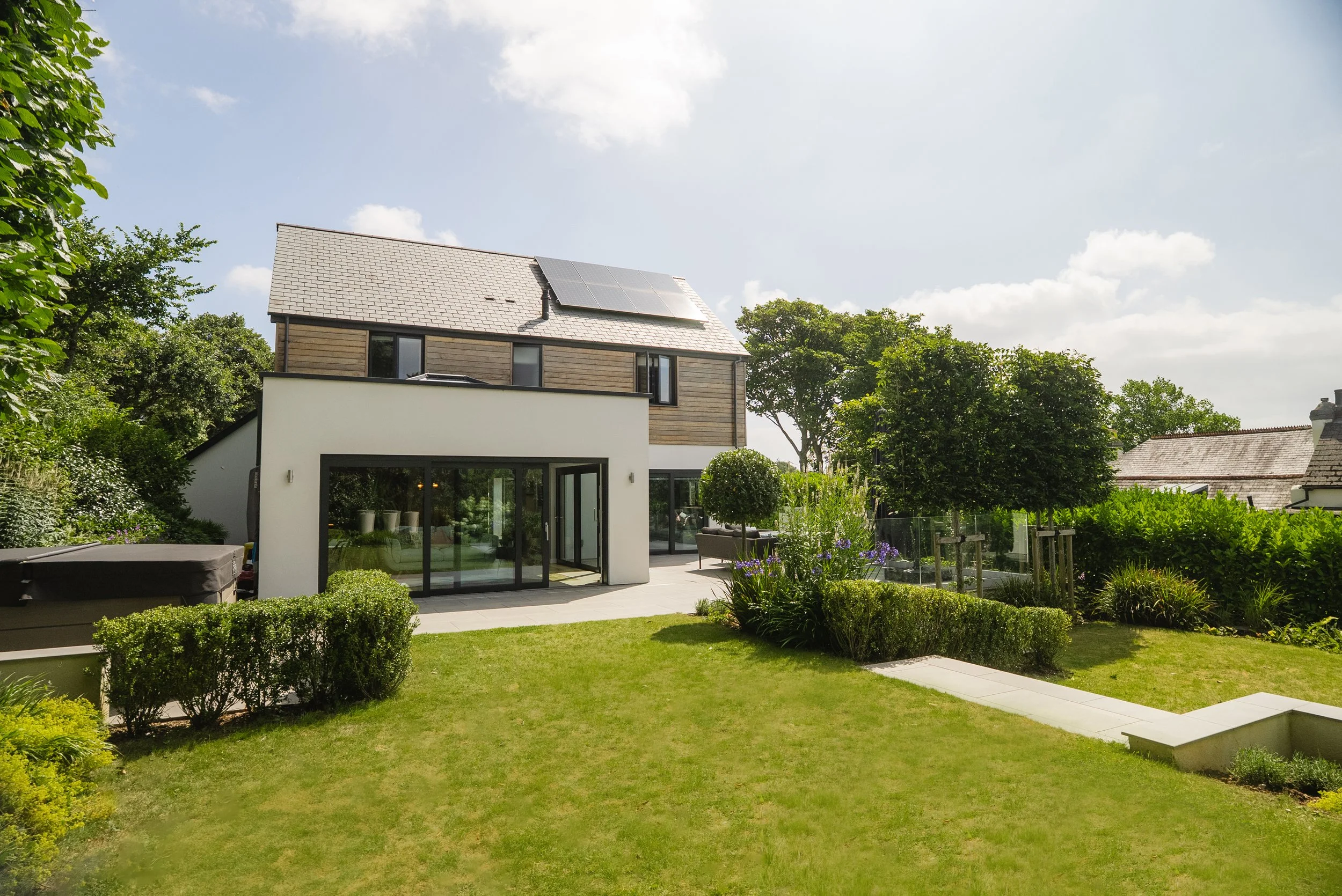 A modern house and garden in Perranwell, Cornwall with large patio doors opening onto a lush lawn bordered by hedges and shrubs and a large seating area and hot tub.