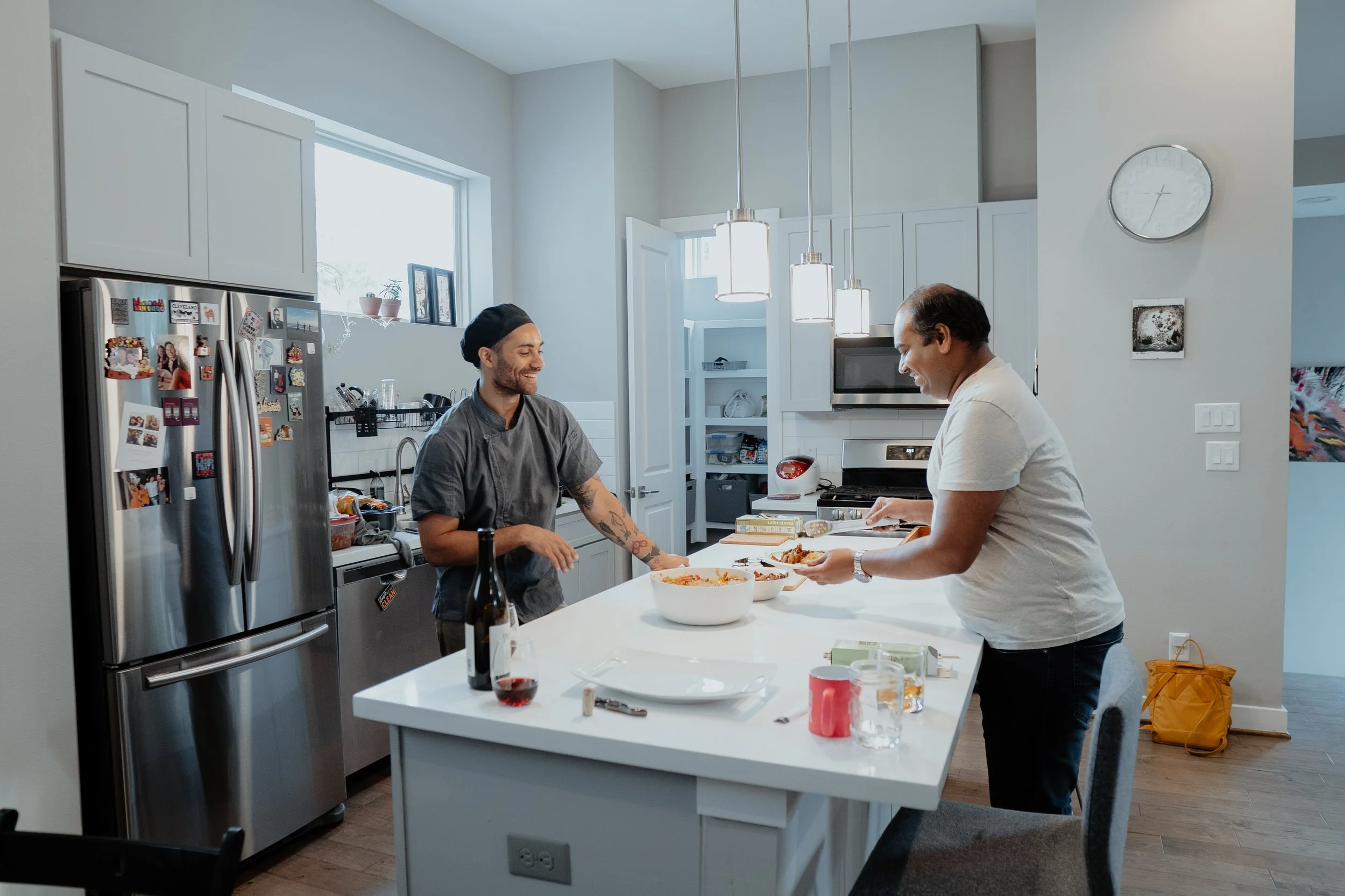 Two men sharing a meal in a modern kitchen, one man is serving food, the other is smiling. The kitchen has white cabinets, a stainless steel refrigerator covered with magnets, and a small window with plants.