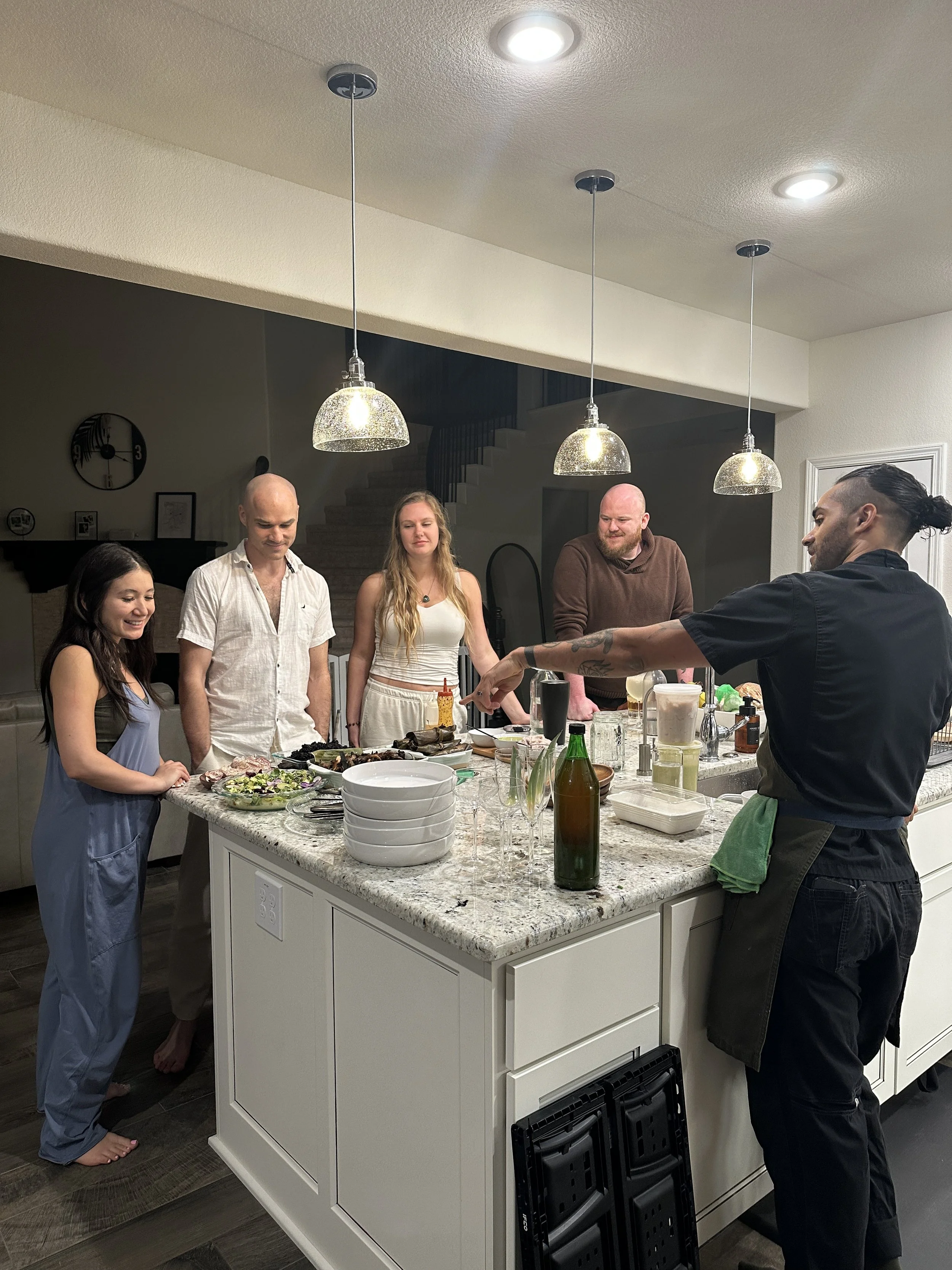 A group of five people gathered around a kitchen island with food and drinks, in a modern home interior with pendant lights and a staircase in the background.