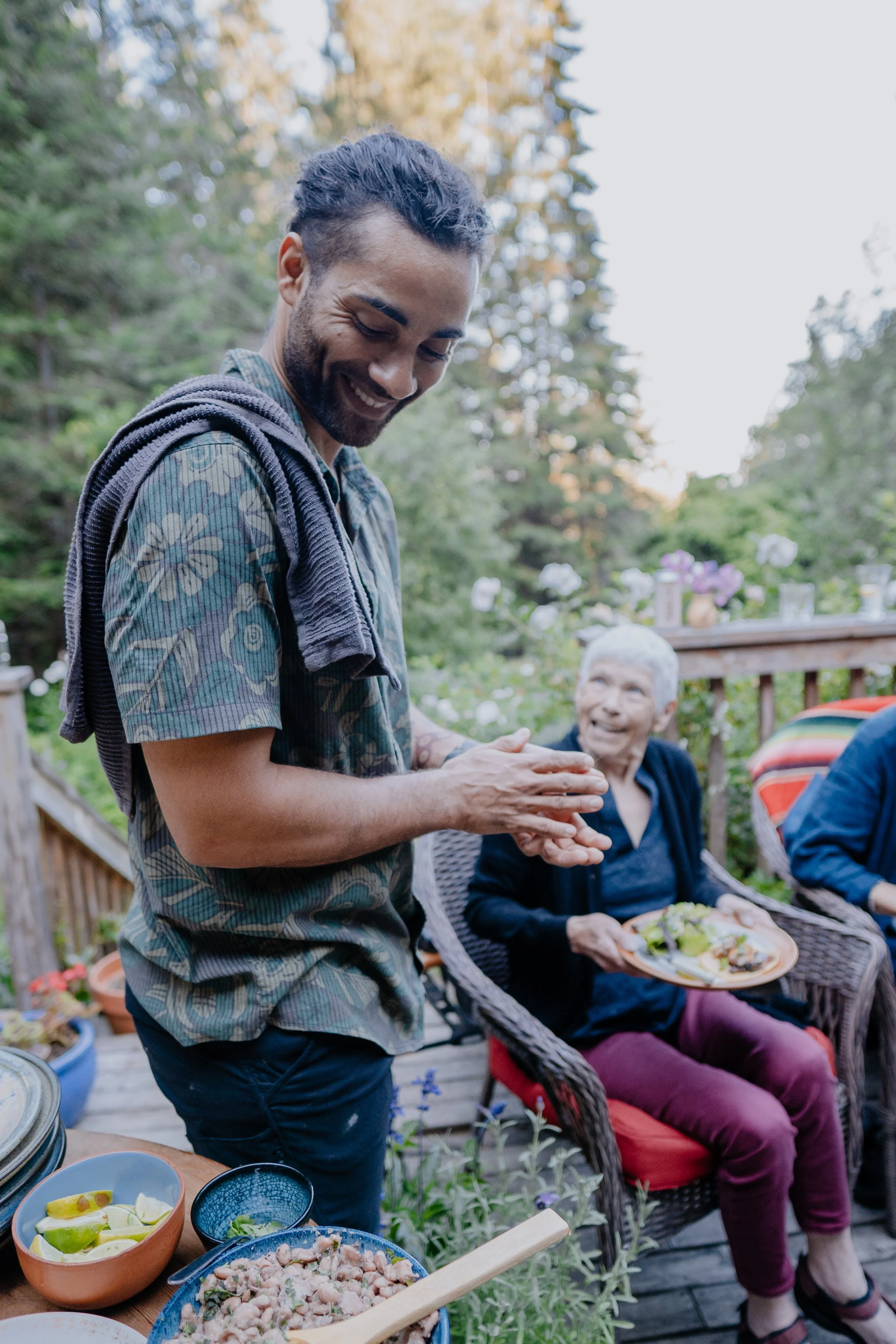 A man with dark hair and a beard is smiling and wearing a patterned shortened-sleeve shirt. An elderly woman with white hair, dressed in dark clothing, is sitting nearby on a red cushioned chair, holding a plate of food. The outdoor setting features a table with various dishes, including bowls and plates of food, and a background of green trees and a wooden deck with string lights and decorative items.