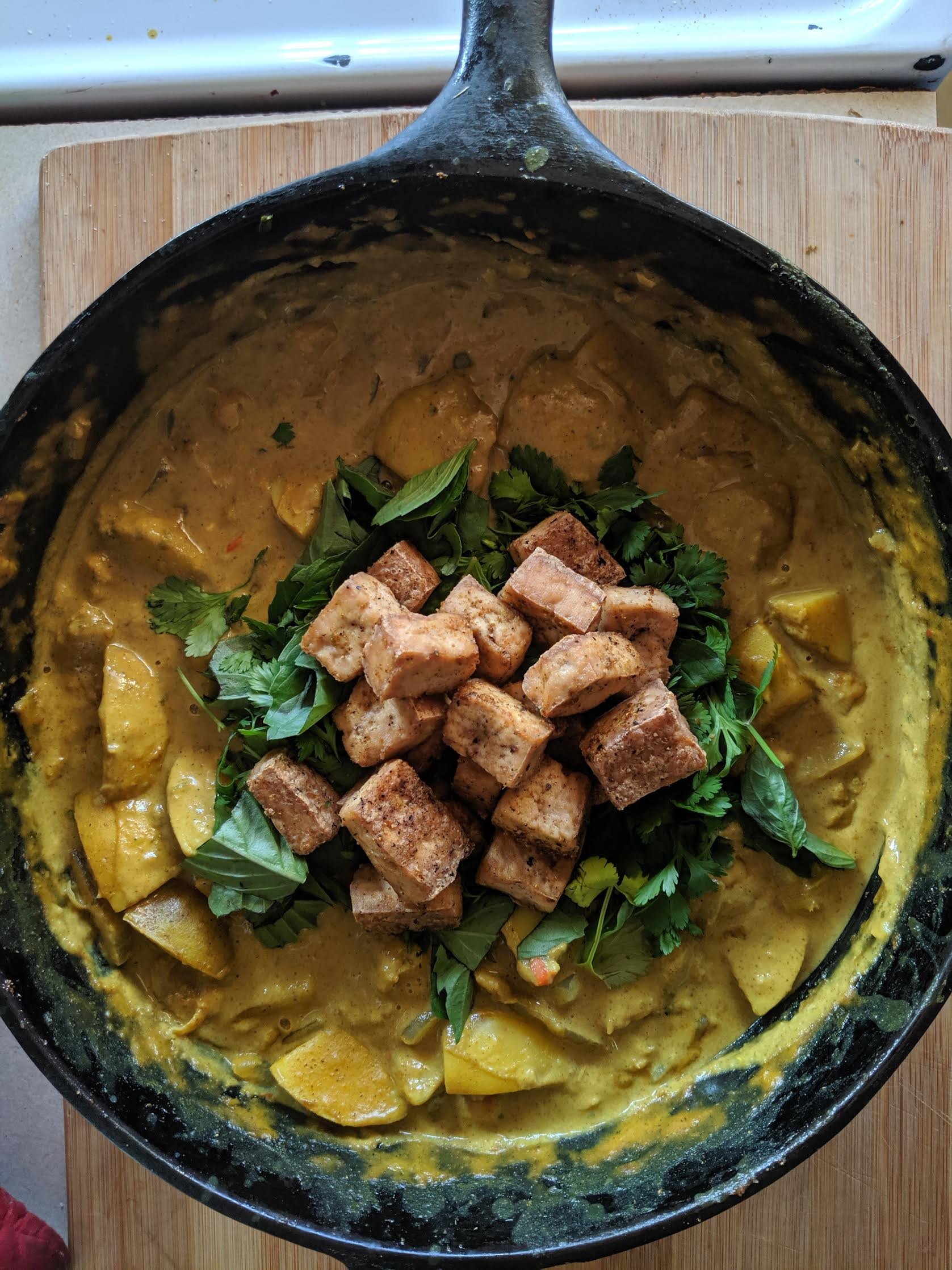 A cast-iron skillet containing a yellow curry dish with cubed tofu and vegetables, garnished with fresh cilantro and basil leaves, placed on a wooden cutting board.