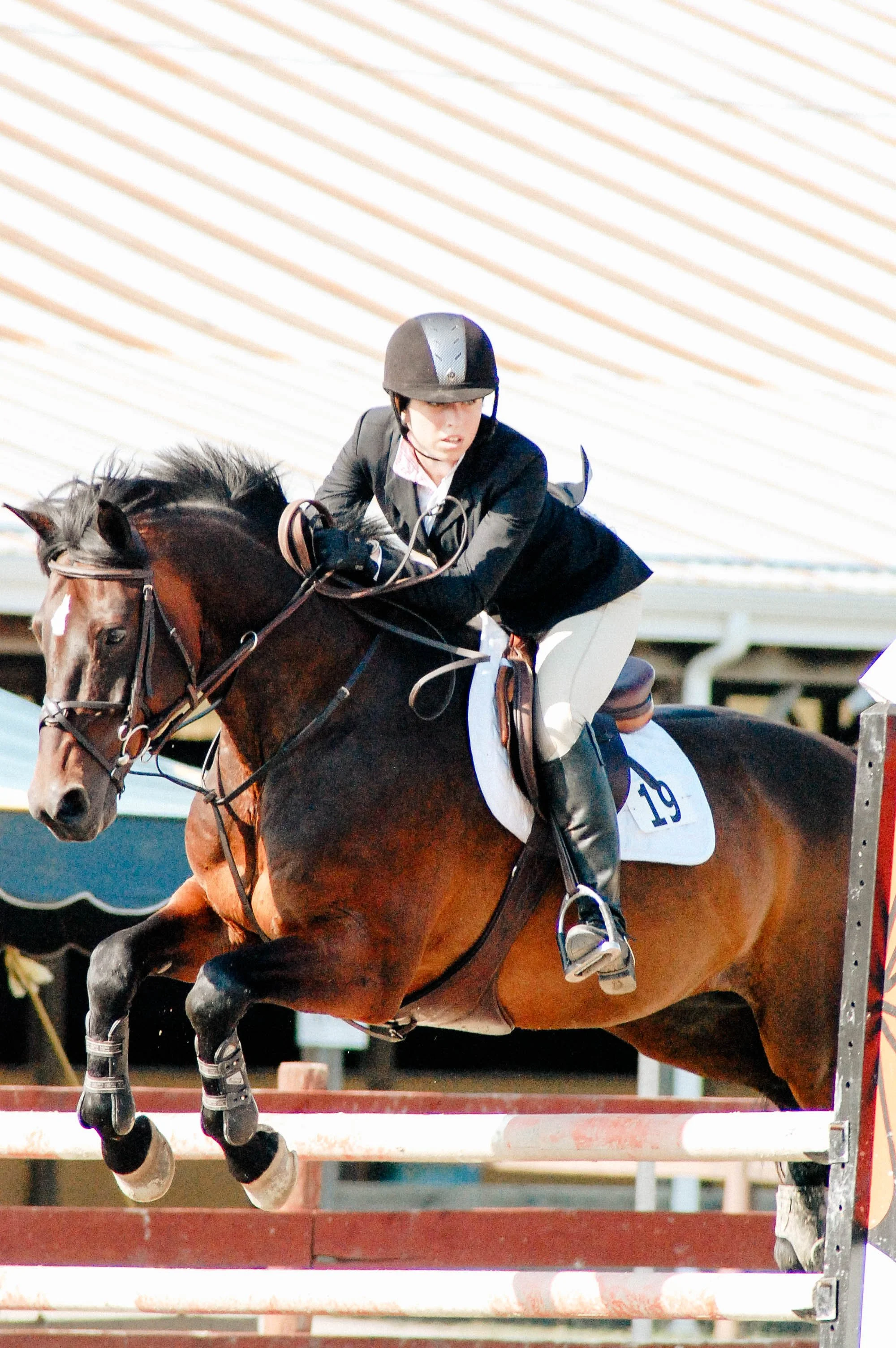 A woman wearing a black helmet and black jacket riding a brown horse over a jump in an equestrian competition.
