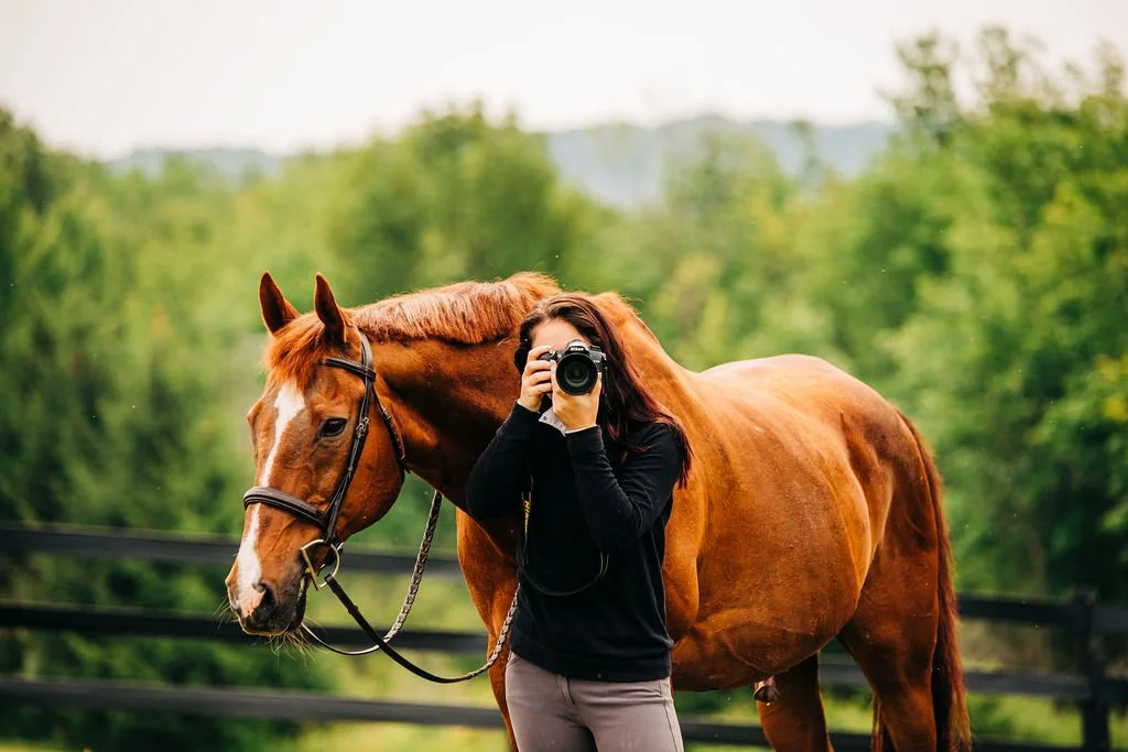 A woman taking a photo of herself with a horse in a green outdoor setting.