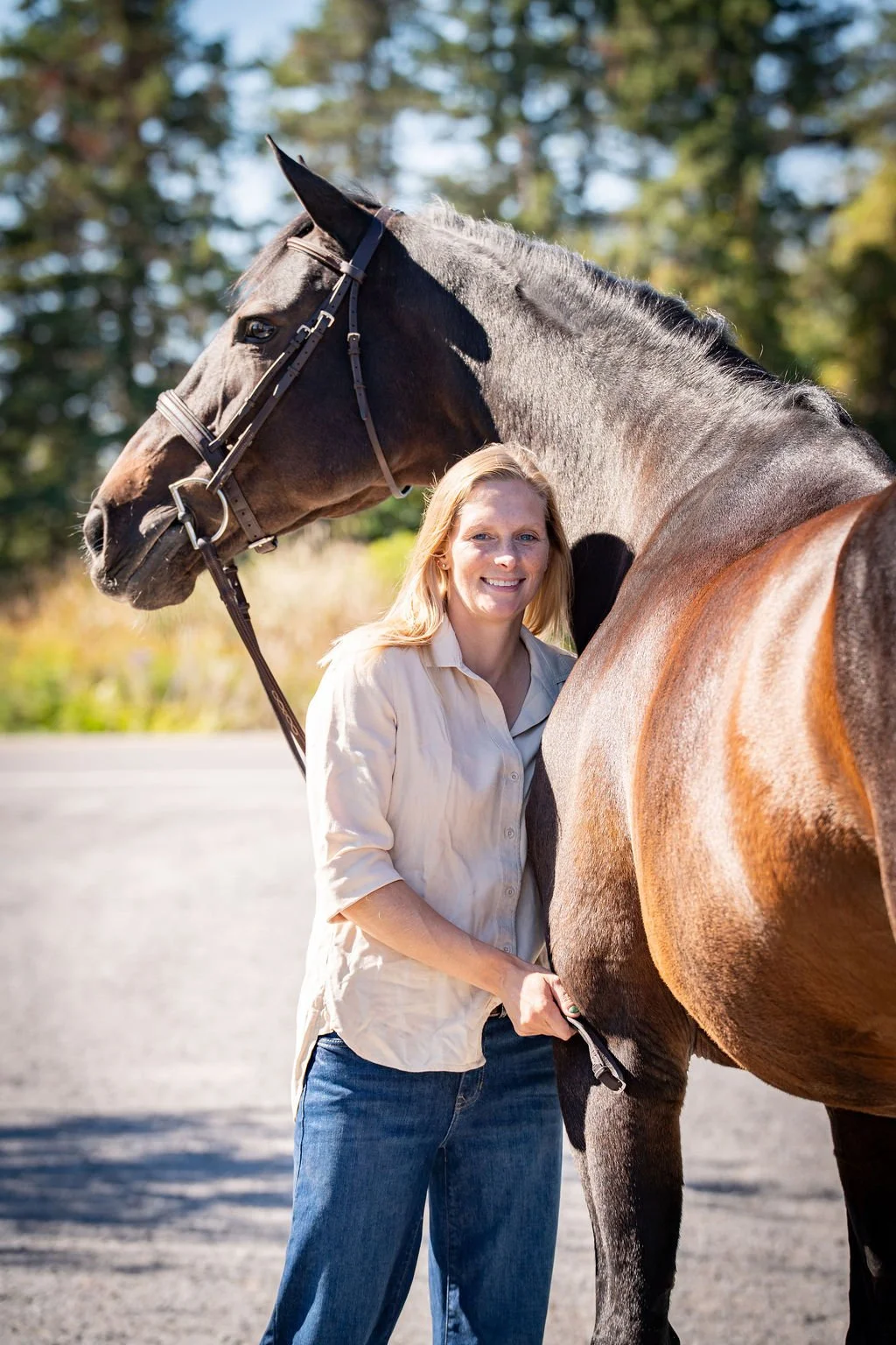 A woman standing next to a brown horse, holding its tail, outdoors on a sunny day with trees in the background.