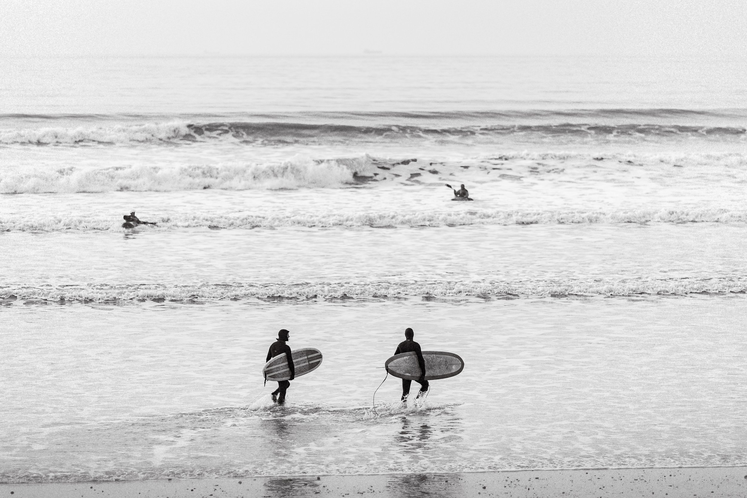 Two surfers walking out of the water holding surfboards at the beach with other surfers riding waves in the background.