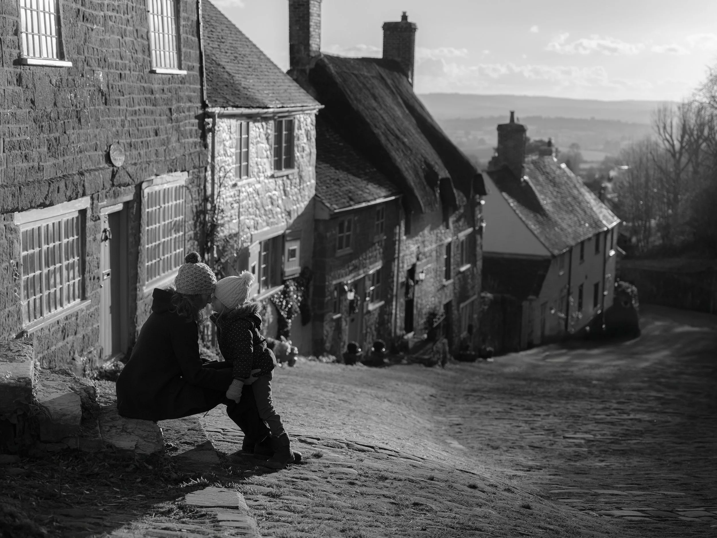 A woman and a littlegirl sitting on a stone ledge in front of a row of historic stone houses with thatched and tiled roofs in a village, with a view of a distant landscape and cloudy sky in black and white. Shaftsbury UK. Fine art photography