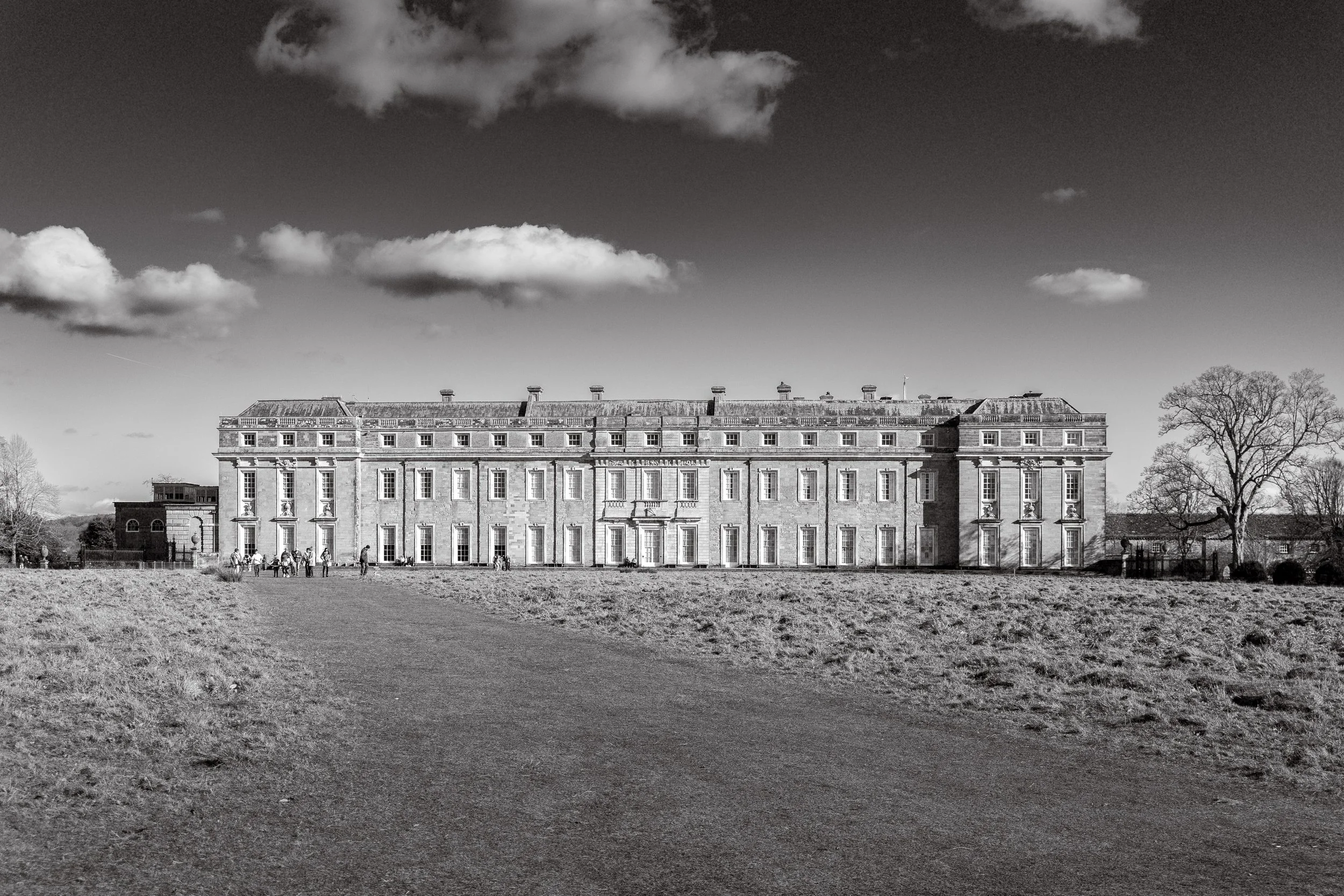 Black and white photograph of a large, historic mansion with a symmetrical facade, multiple windows, and a central entrance, set against a partly cloudy sky with trees on either side.