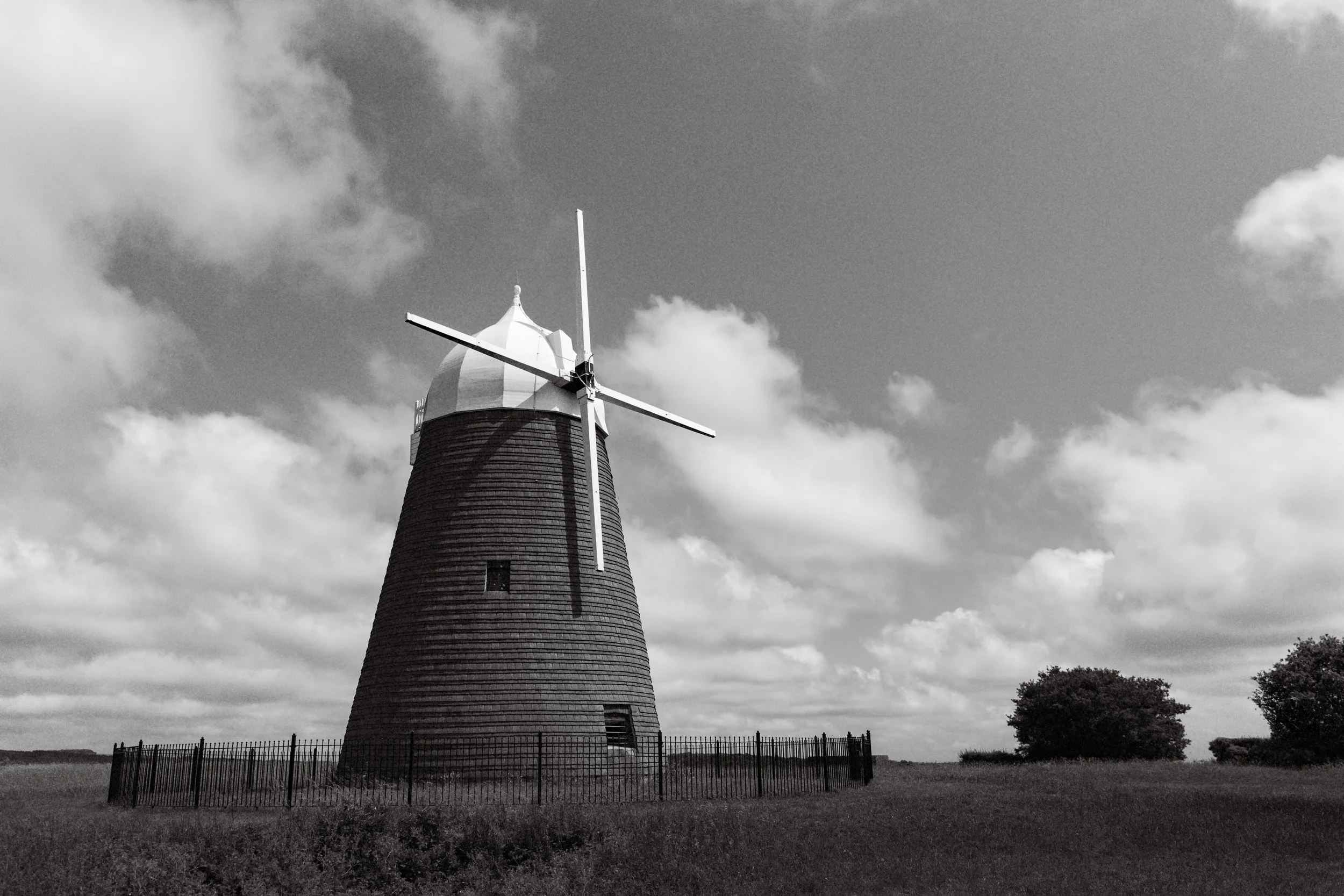 A windmill with a dark, textured body and white blades stands in an open field under a partly cloudy sky. Fine Art Landscape Photography