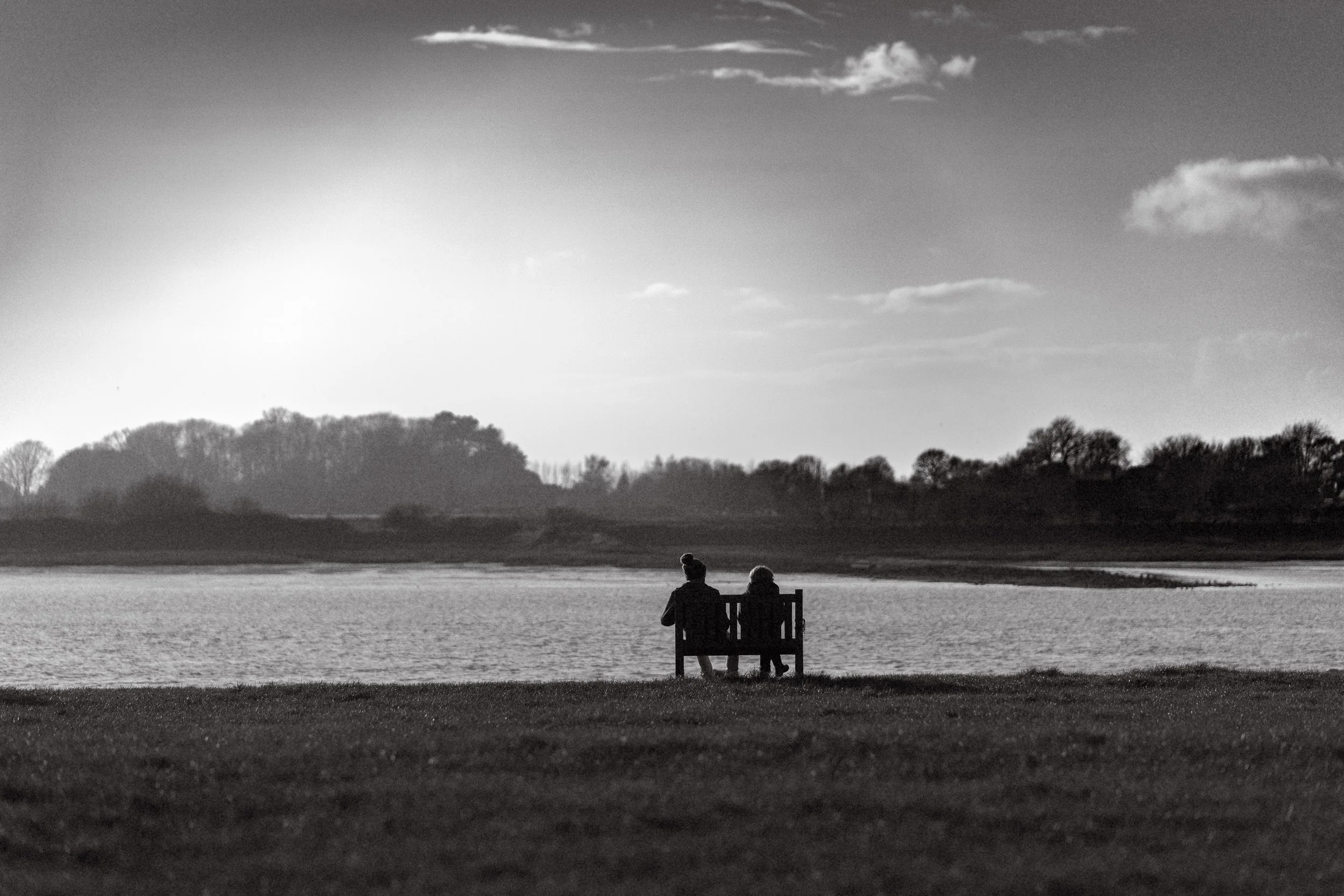 Two people sitting on a bench facing a body of water, with trees in the background and a few clouds in the sky, in black and white.