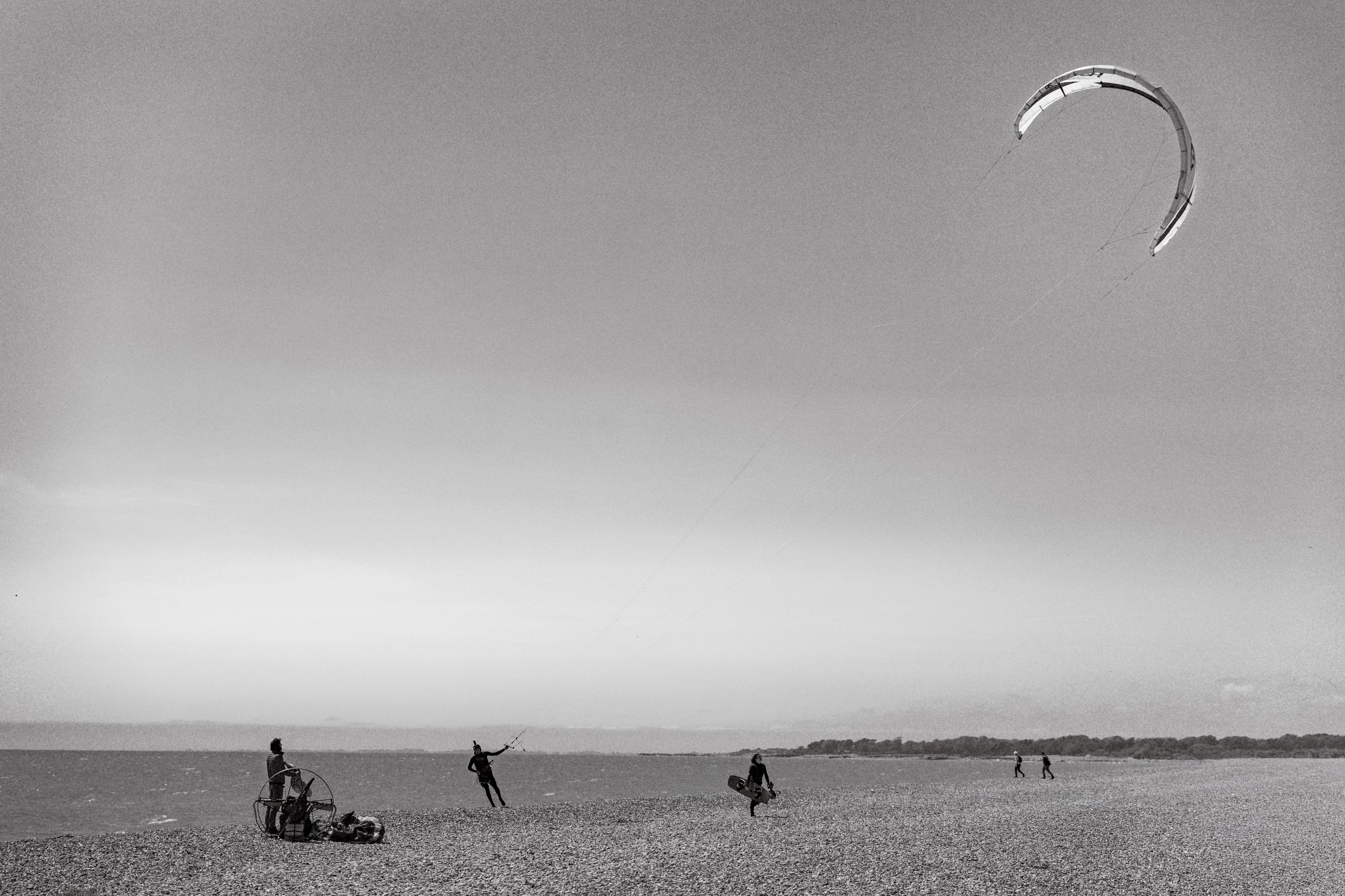 People on a beach flying a large kite in the sky, some walking, some sitting, with a distant shoreline.