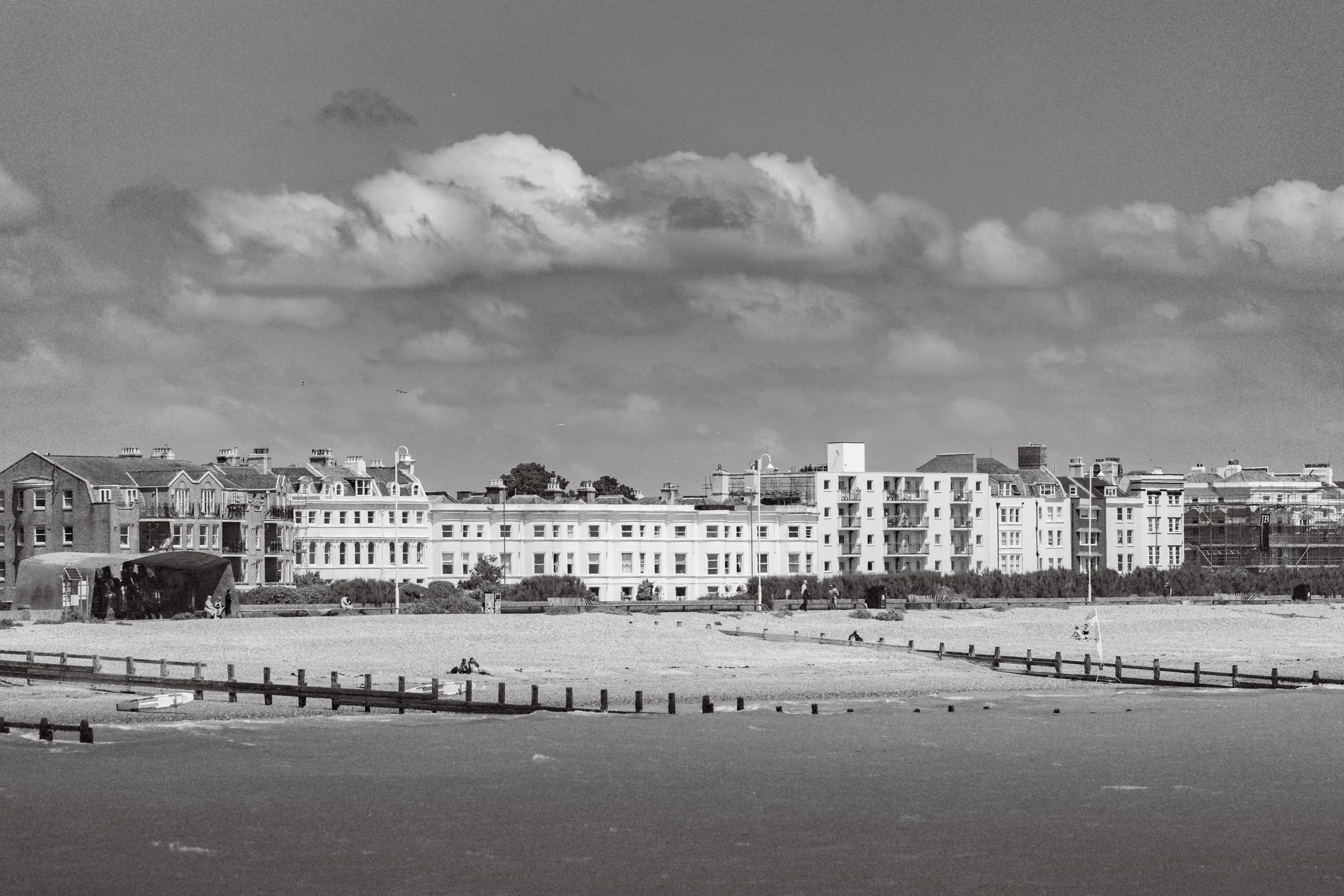 A black and white photo of a beach with a sandy shore, small boats, and a wooden fence. In the background, there are several multi-story residential buildings under a cloudy sky.