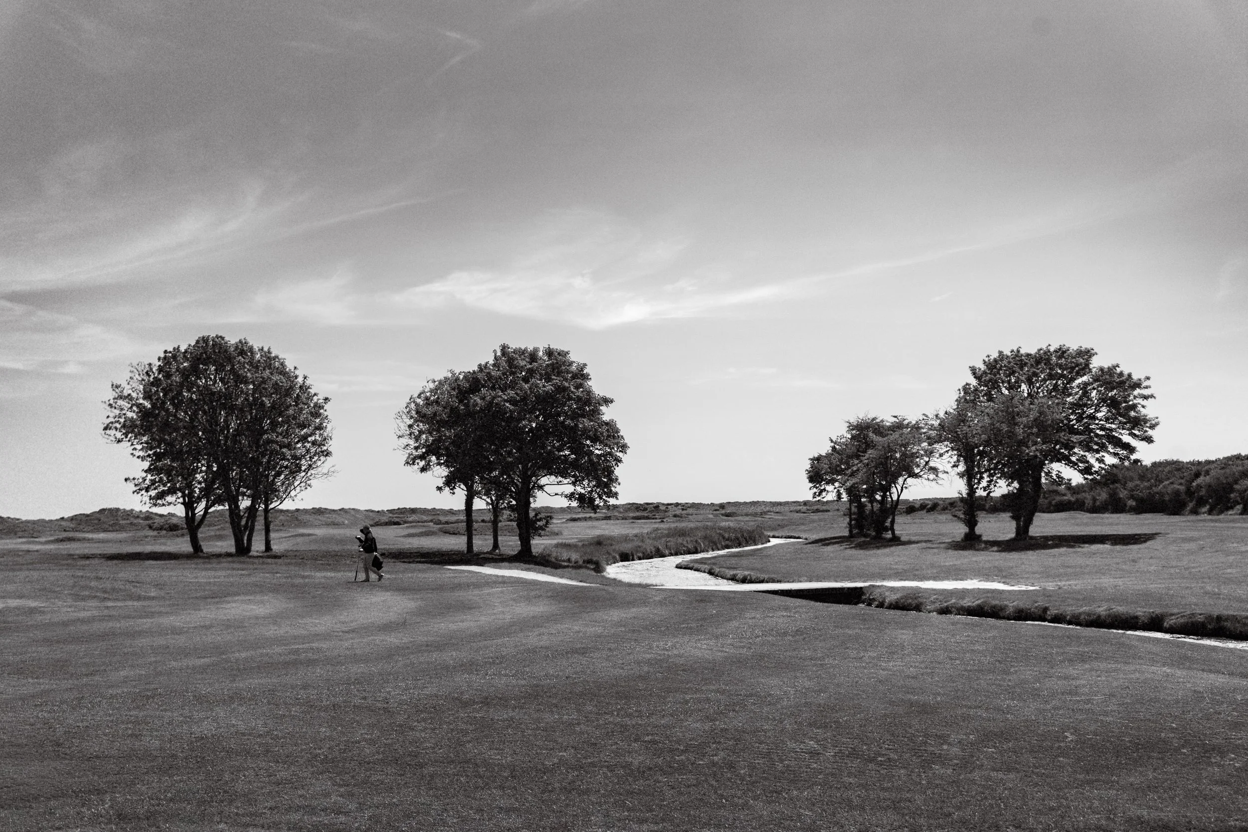 A golf course with a golfer walking near trees and a small water hazard, under a partly cloudy sky. Fine Art Landscape Photography
