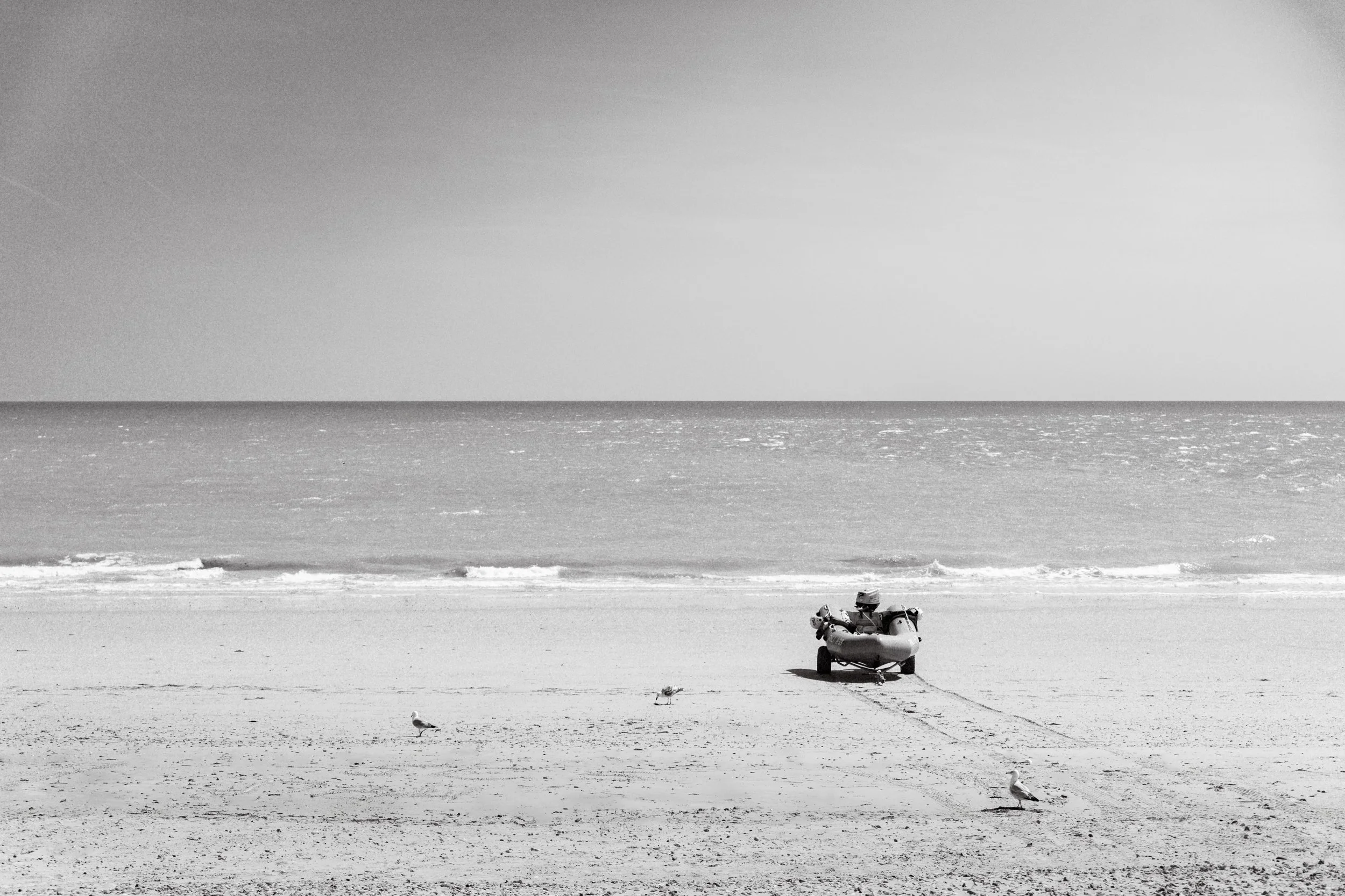 Black and white photo of a serene beach with a small boat on the sand, seagulls, and calm ocean waves under a clear sky. Fine Art Landscape Photography