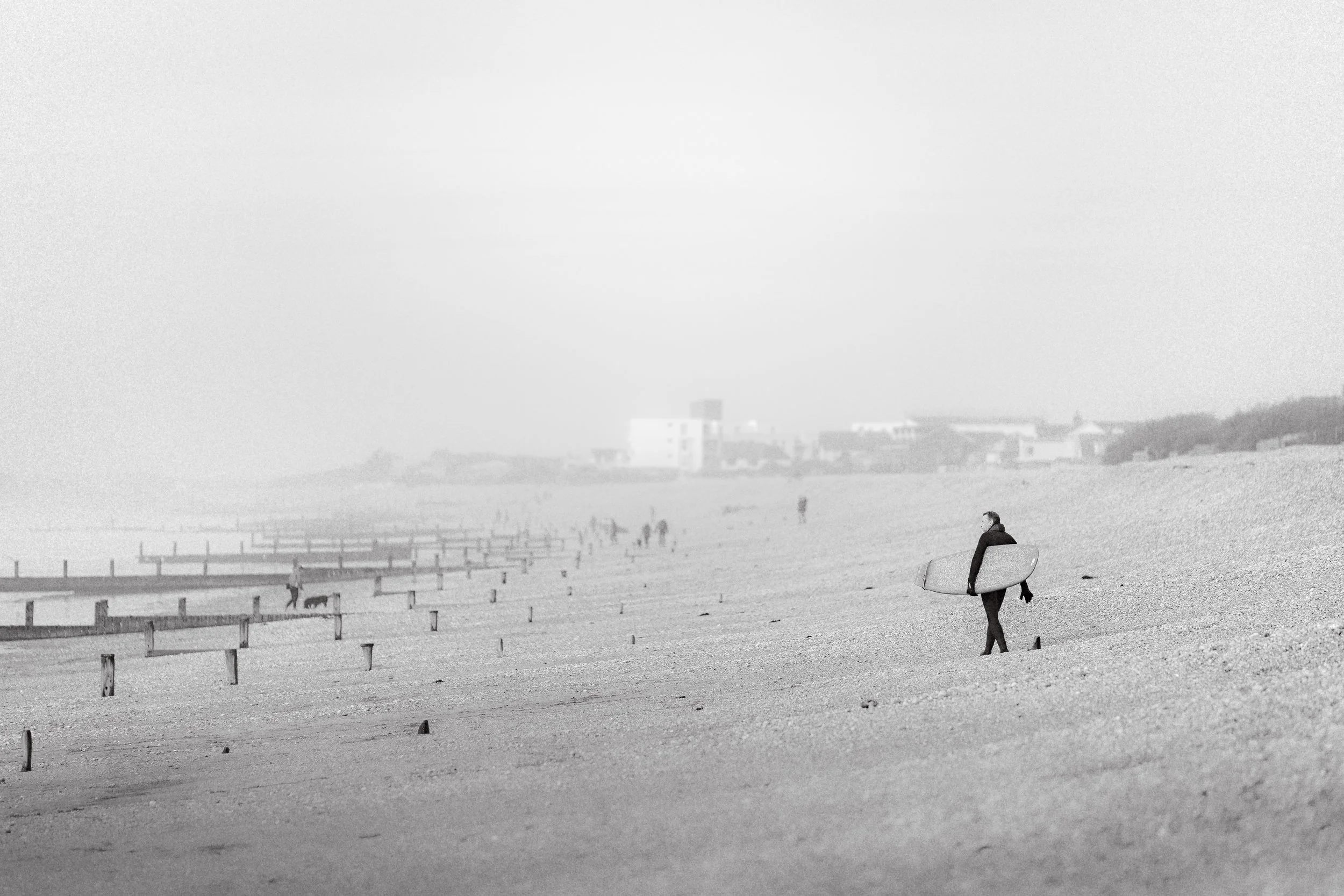 A person with a surfboard walking along a deserted beach, with a hazy background of other people, buildings, and a fence.