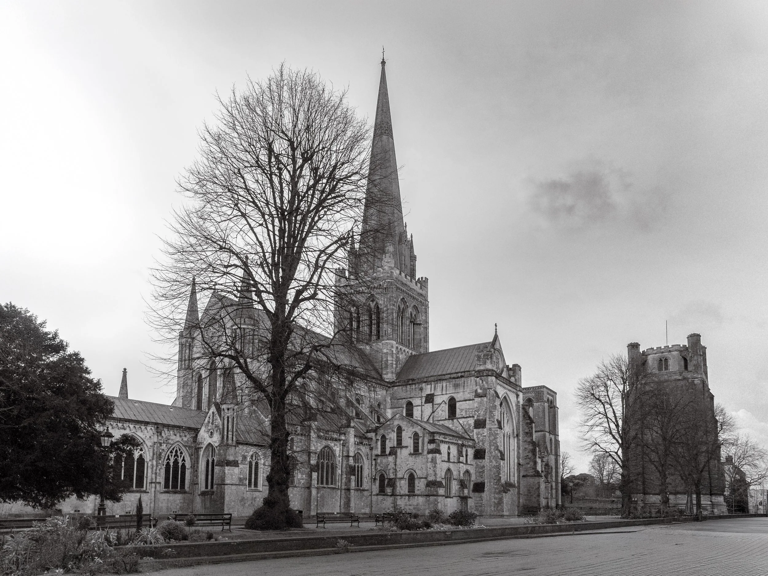 Black and white photo of a large Gothic church with tall spires, surrounded by leafless trees and benches in the foreground.