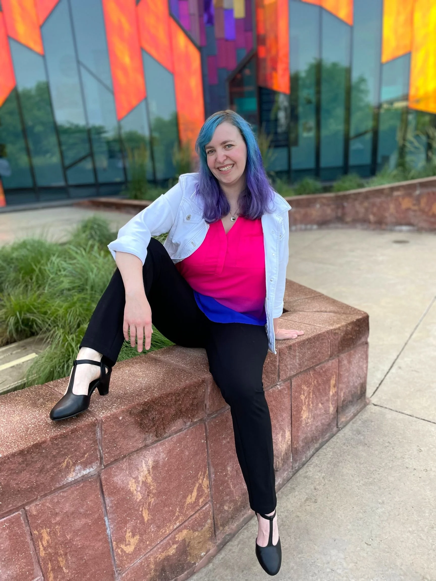 Woman with blue and purple hair wearing a white jacket, pink shirt, and black pants and shoes is perched on a wall with a slight lean. She is smiling and sitting in front of colored glass on a building behind her.