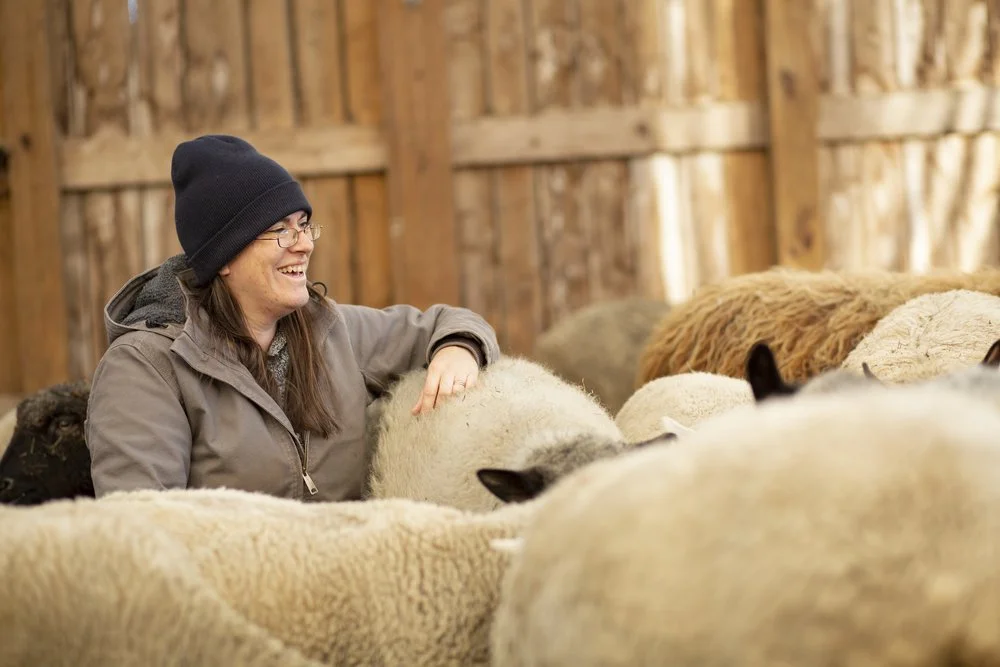 Azsa Greiner stands among sheep inside of a wooden structure wearing a winter hat and coat