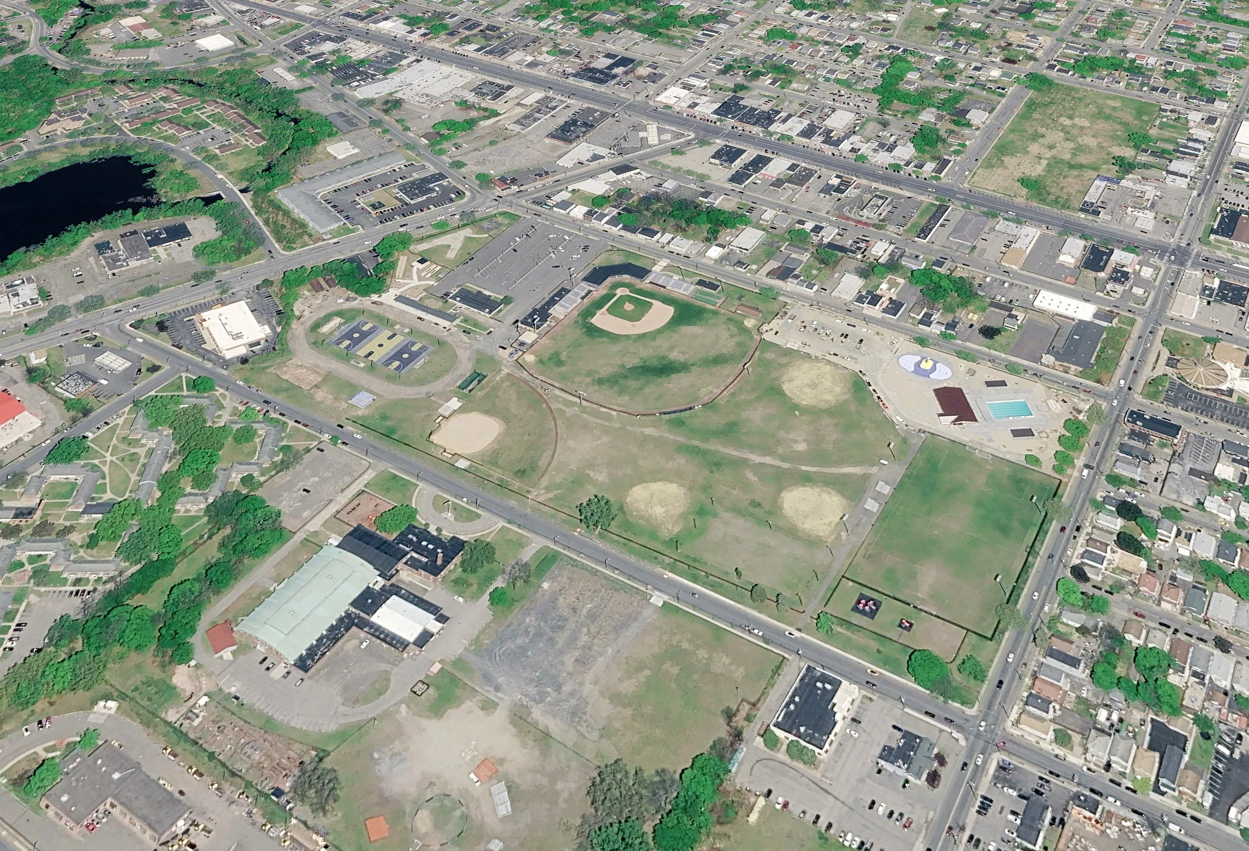 An aerial view of a city park with baseball fields, tennis courts, playgrounds, and nearby residential and commercial buildings. Streets and green trees surround the park.