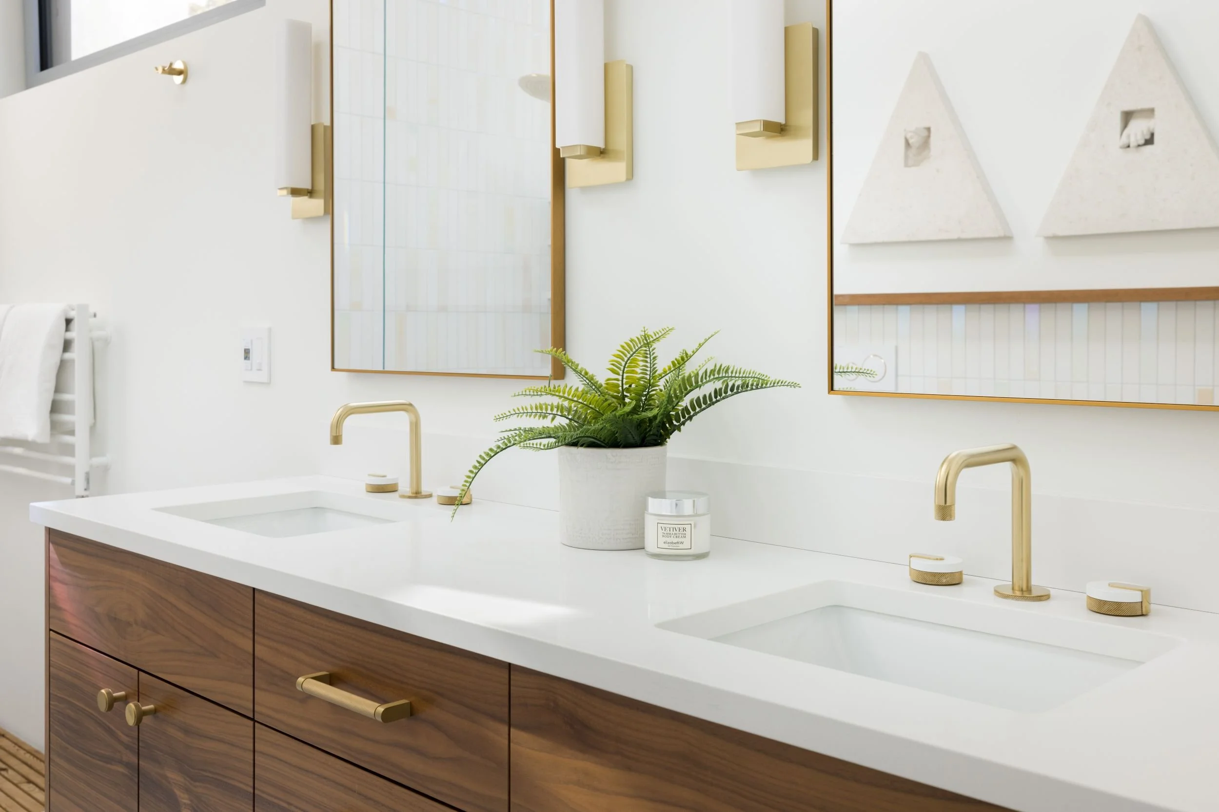 Modern bathroom with a double vanity, gold fixtures, wood cabinets, large mirror, potted fern, candle, and decorative art on the wall.