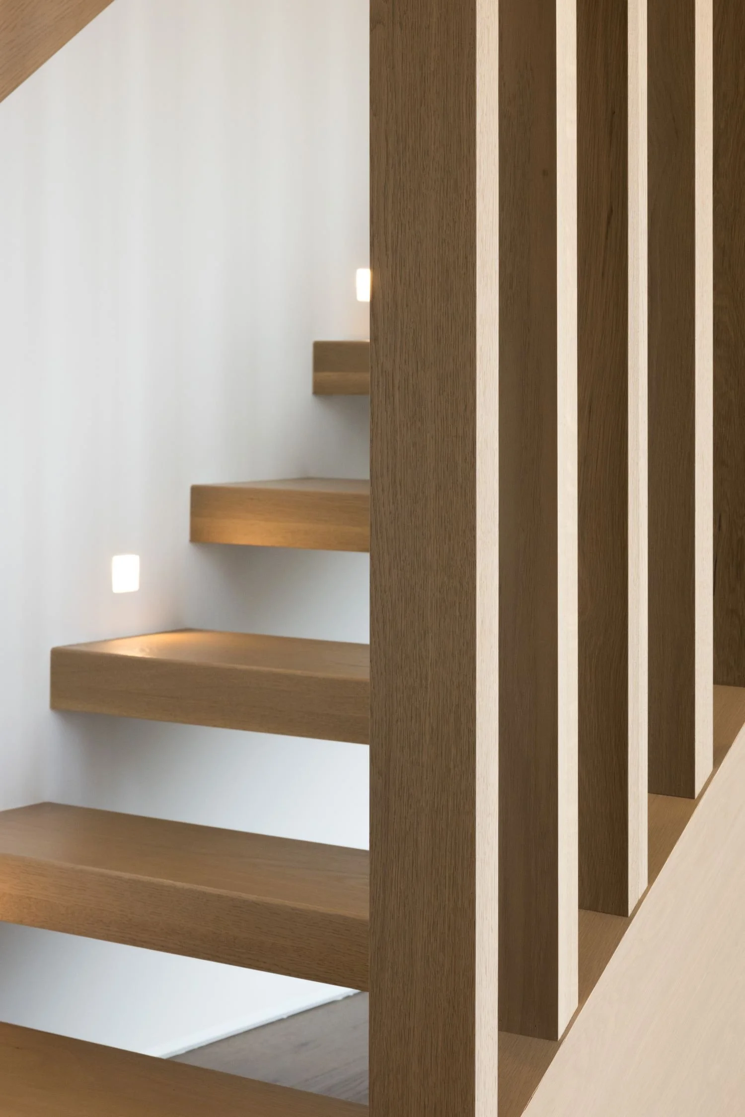 Close-up of a modern, minimalist wooden staircase with floating steps and vertical wooden slats, illuminated by small square lights in a contemporary interior.